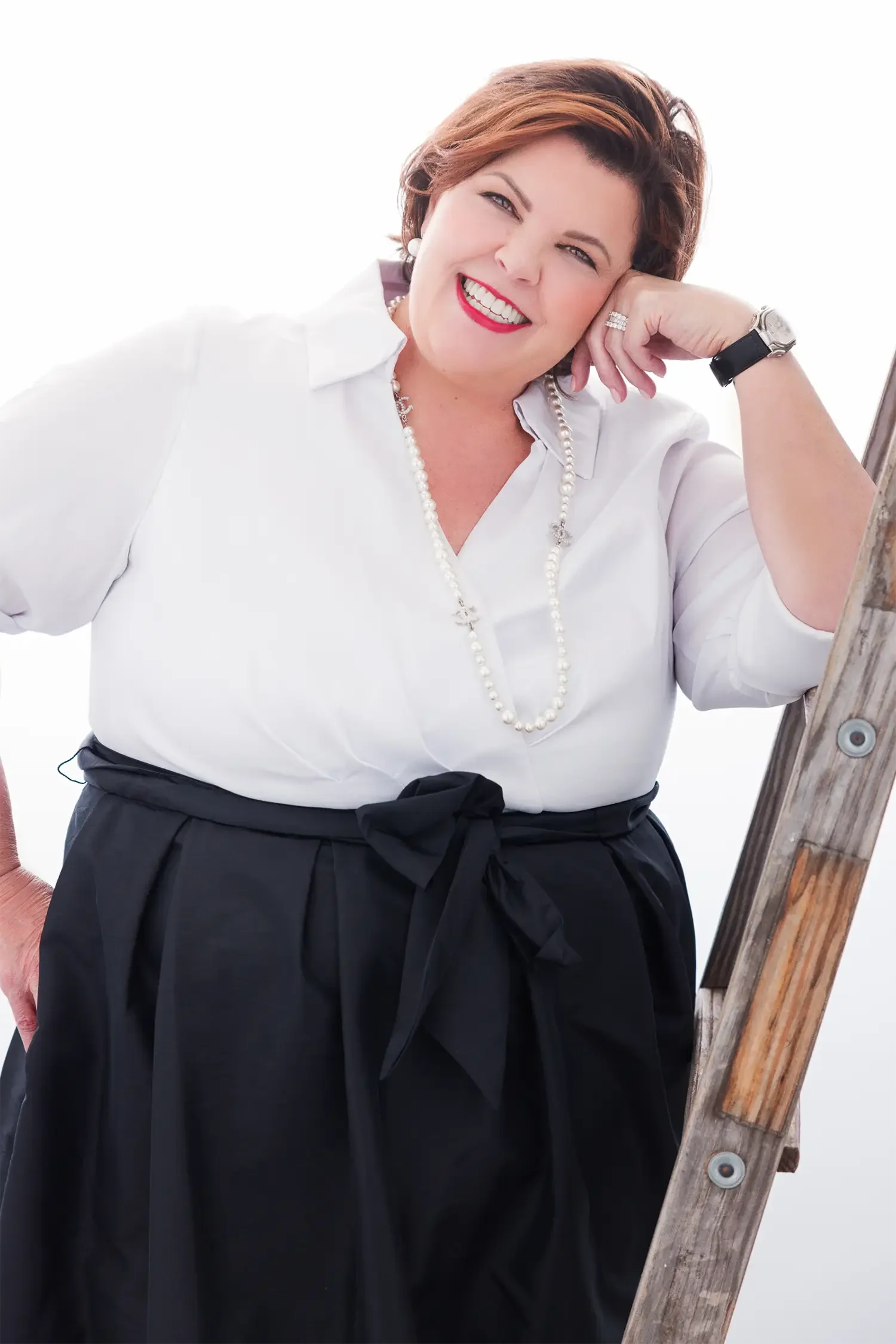 A woman smiling, leaning on a wooden ladder, dressed in a white blouse and black skirt with a bow, wearing pearl jewelry and a watch, against a white background.