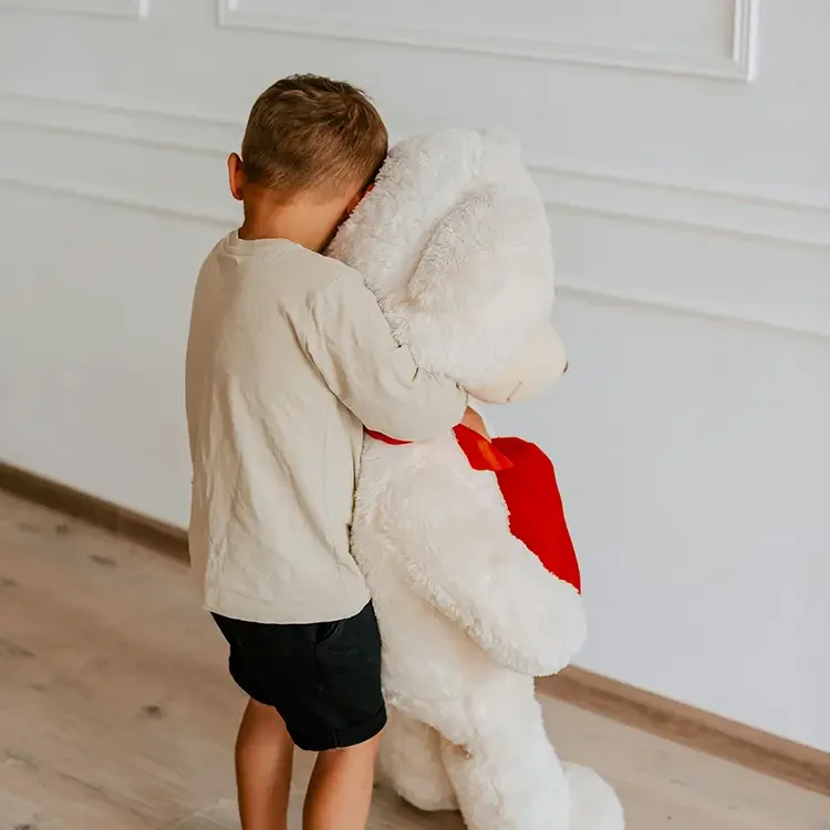 A young boy hugging a large white teddy bear with a red ribbon, inside a room with wooden flooring and white paneled walls.