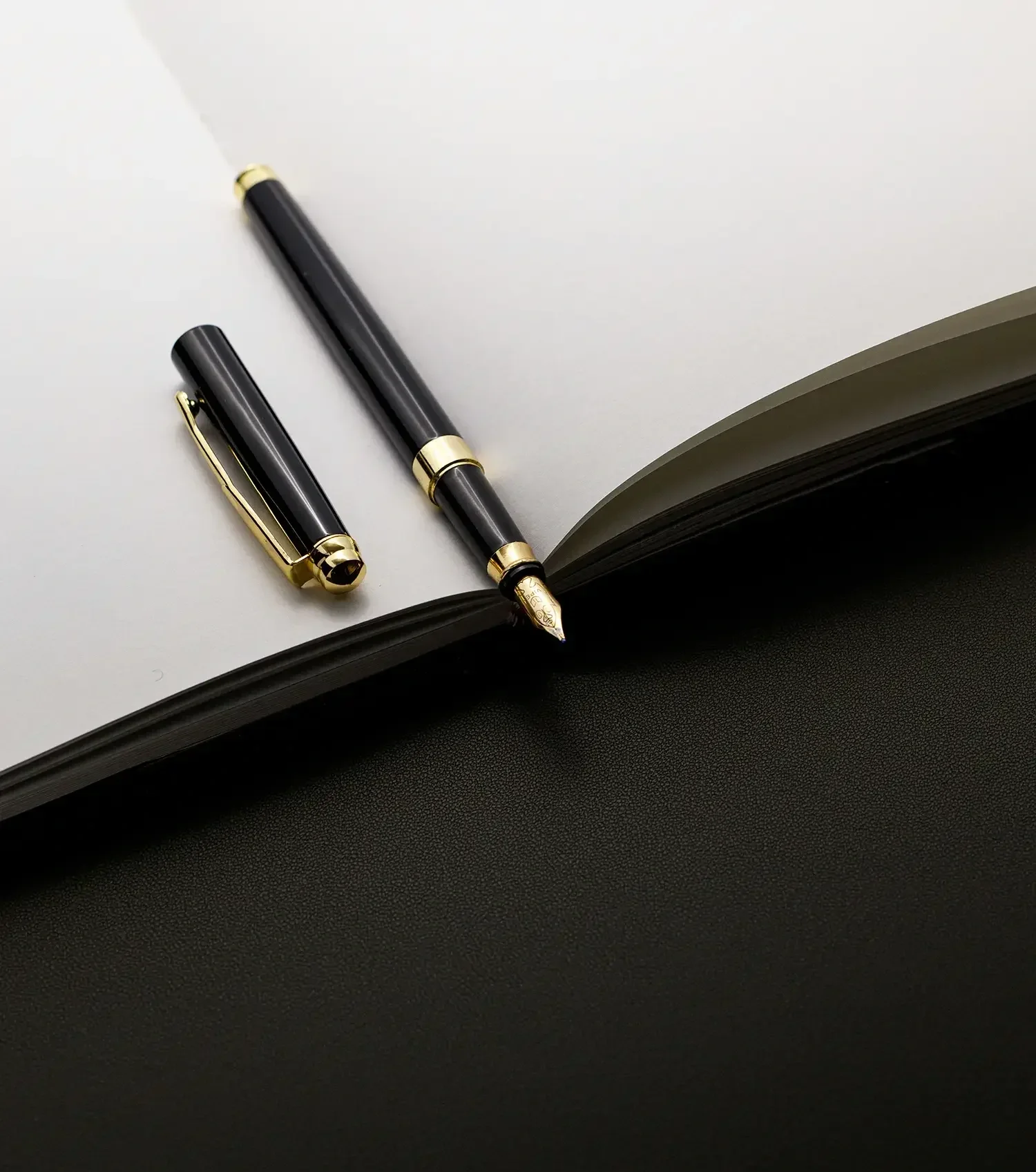 Black fountain pen with gold accents resting on an open blank notebook with a black cover, on a white surface.