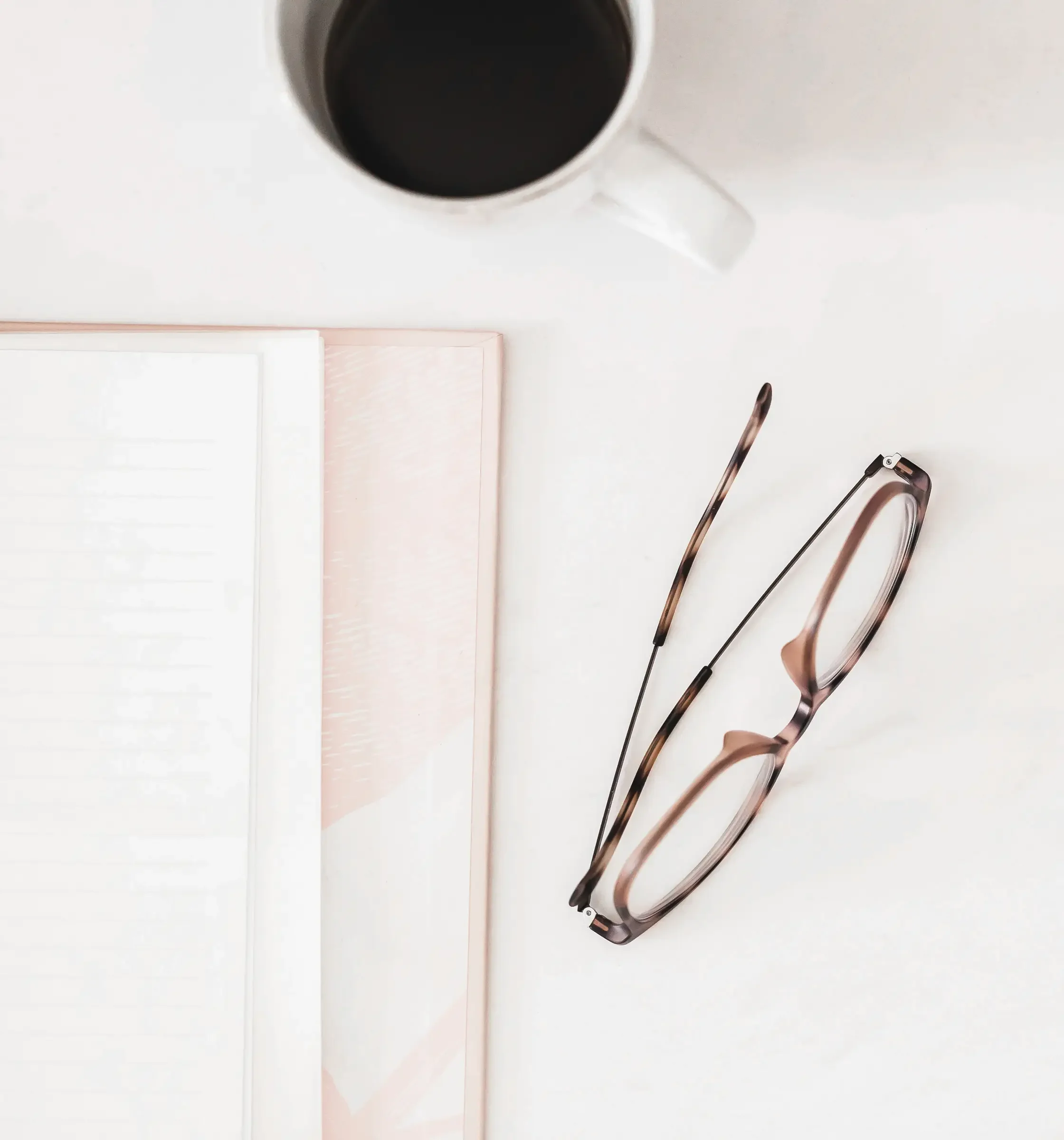 A top-down view of a white workspace with a cup of black coffee, a pink notebook, and eyeglasses with tortoise shell frames.