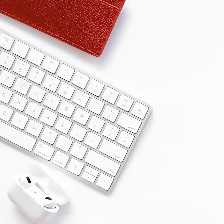 Wireless earbuds with charging case, white keyboard, and a red textured notebook on a white surface.