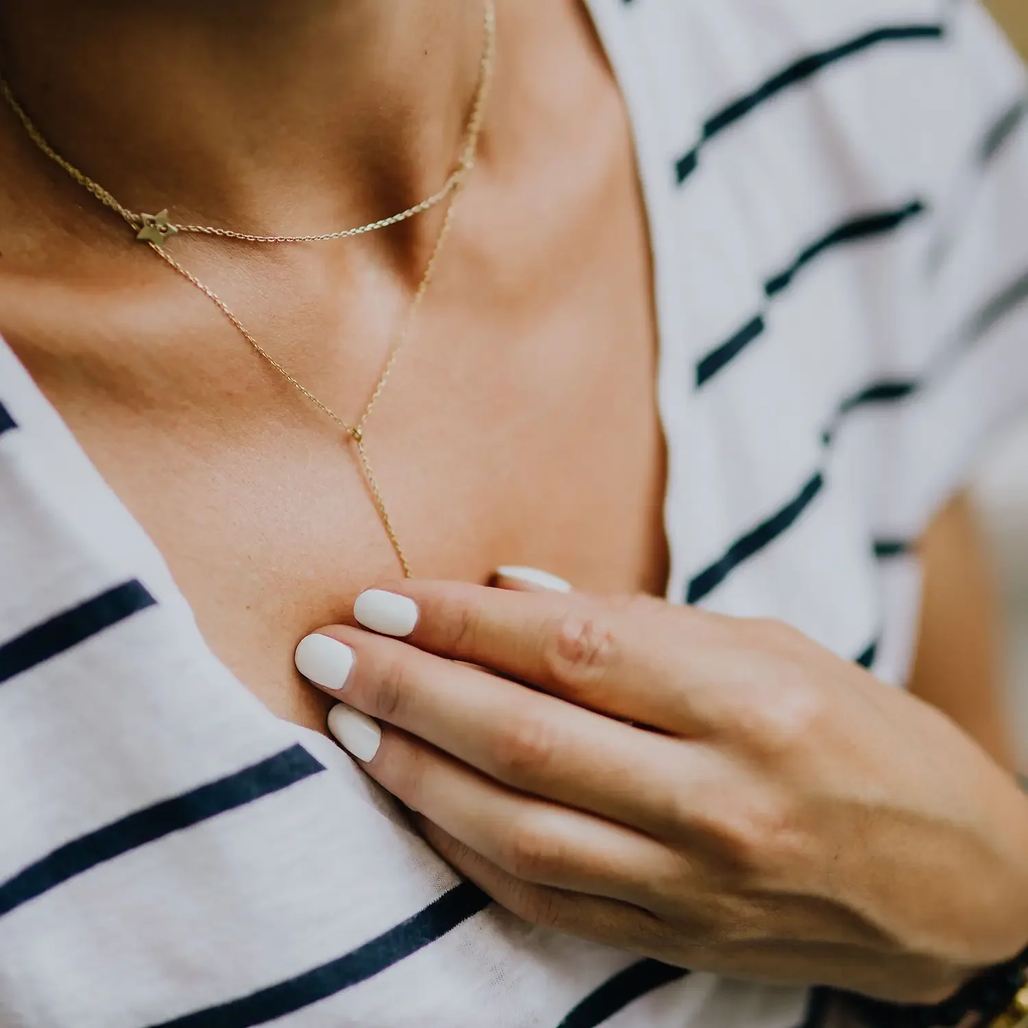 Close-up of a woman wearing layered gold necklaces, with one hand resting on her chest, wearing a striped shirt with navy and white lines.