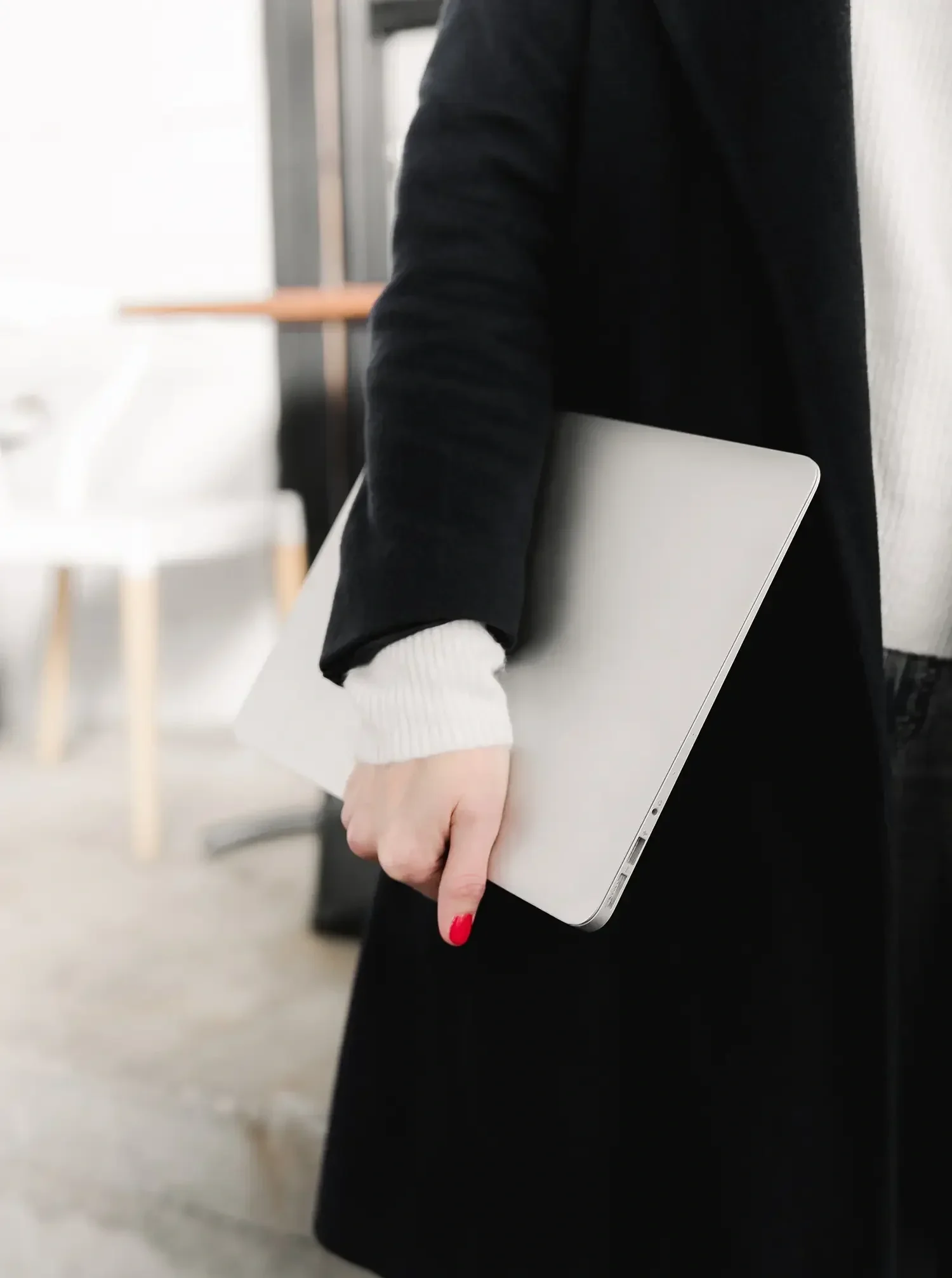 Person holding a closed silver laptop, dressed in a black coat with a white sleeve and red nail polish, in an indoor setting.