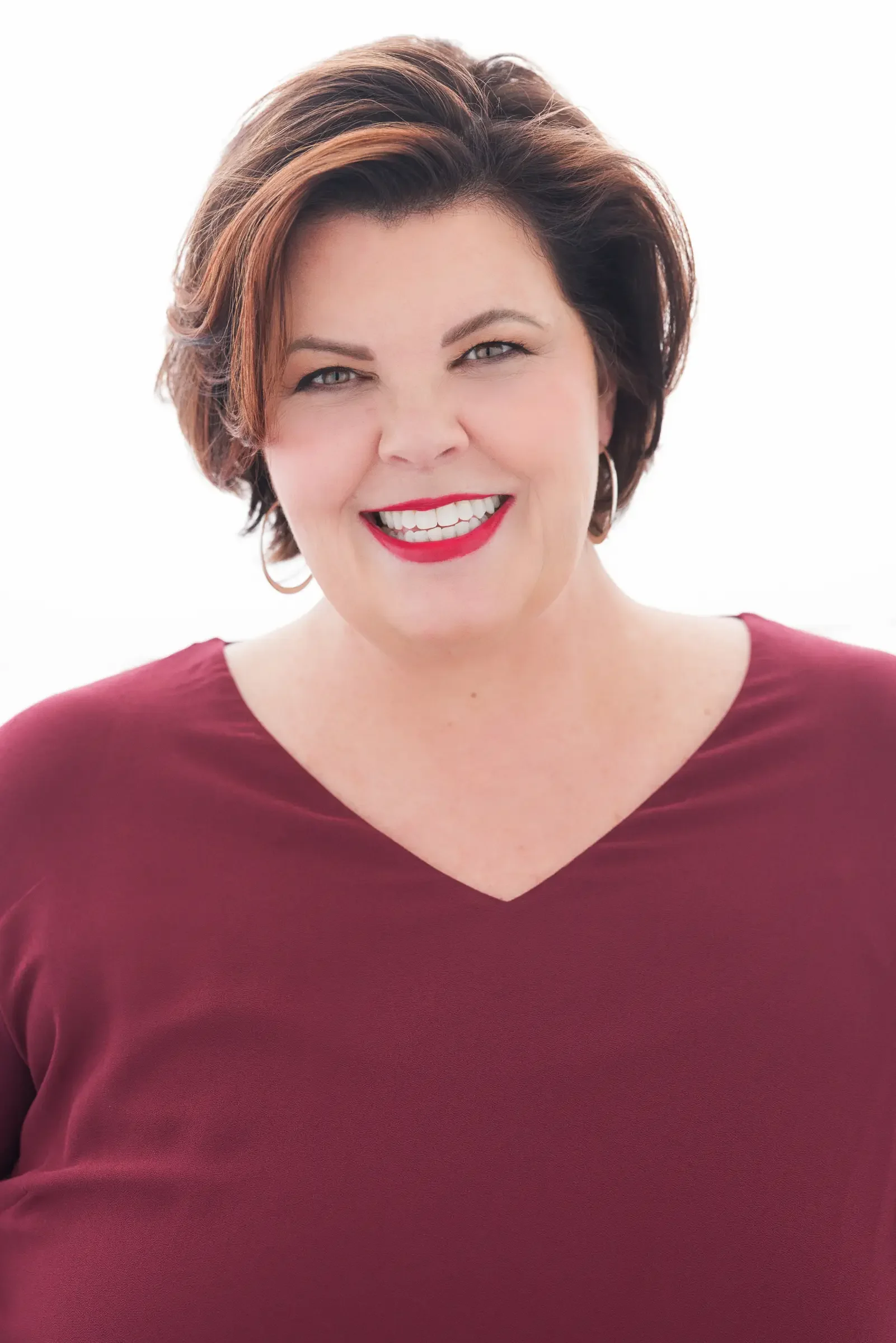 A woman with short dark hair, wearing a maroon top, smiling and showing her teeth, with bright lipstick and hoop earrings.