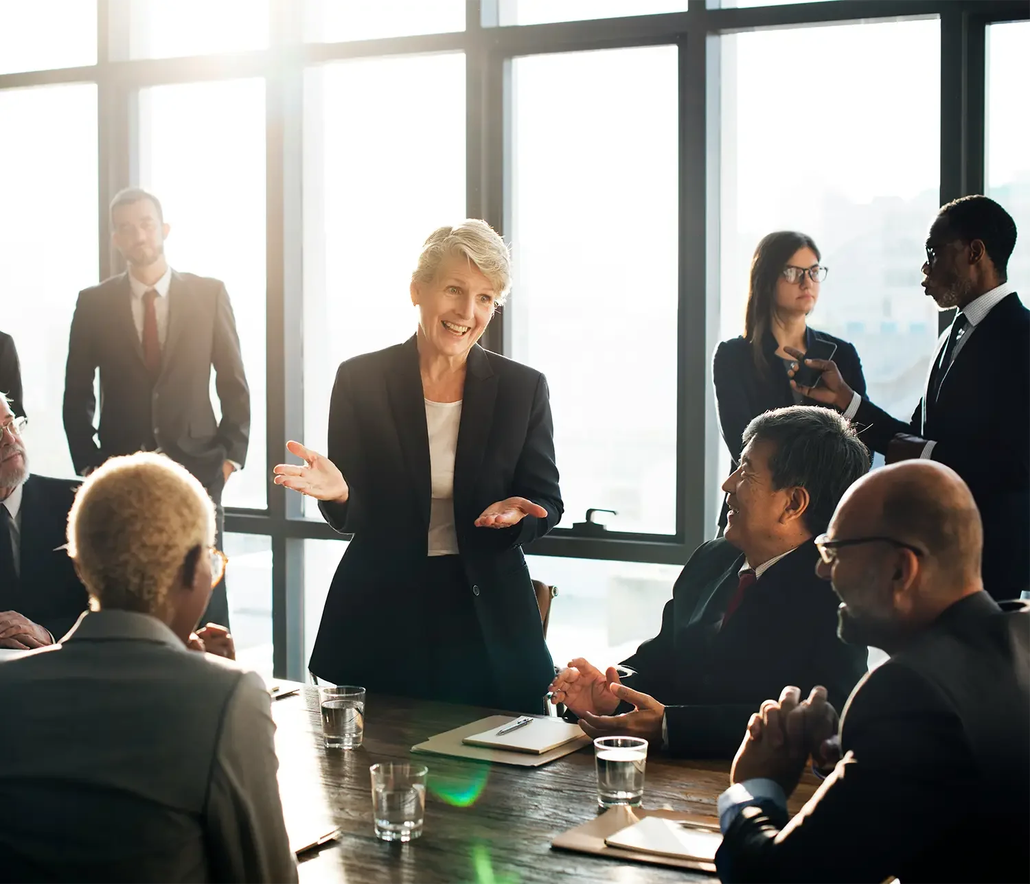 Business meeting with a woman speaking and smiling, while seated and surrounded by professionals in a conference room with large windows.