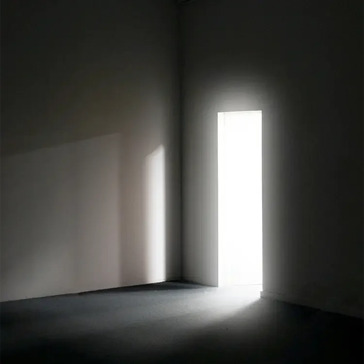 An empty room with a tall, narrow door letting in bright natural light, creating shadows on the walls and floor.
