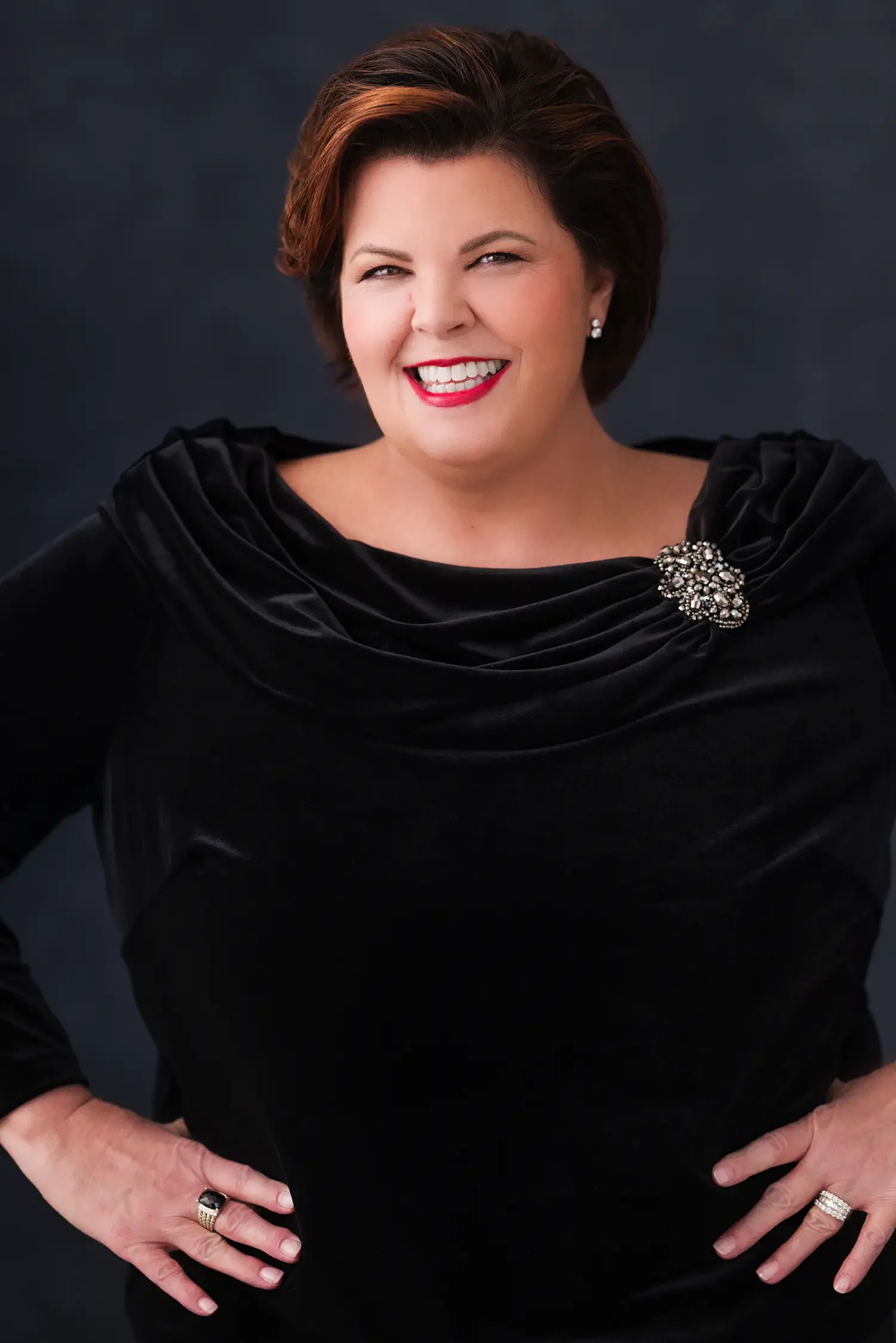 A smiling woman with short, reddish-brown hair wearing a black velvet dress with a brooch, standing against a dark background.
