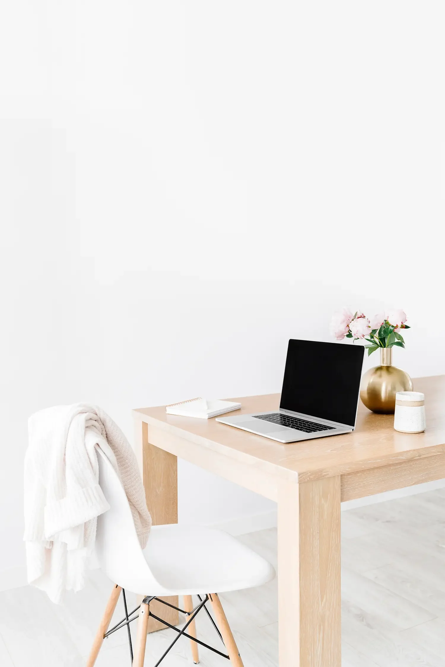 Minimalist workspace with a light wood desk, a white chair draped with a blanket, a laptop, a small notebook, a gold vase with pink flowers, and a small decorative container, set against a plain white wall.