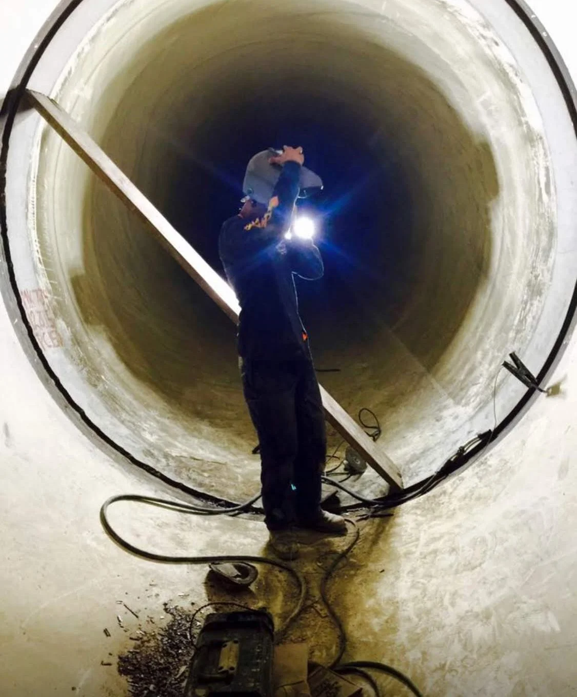Worker welding inside a large pipe, with tools and cables on the ground nearby.