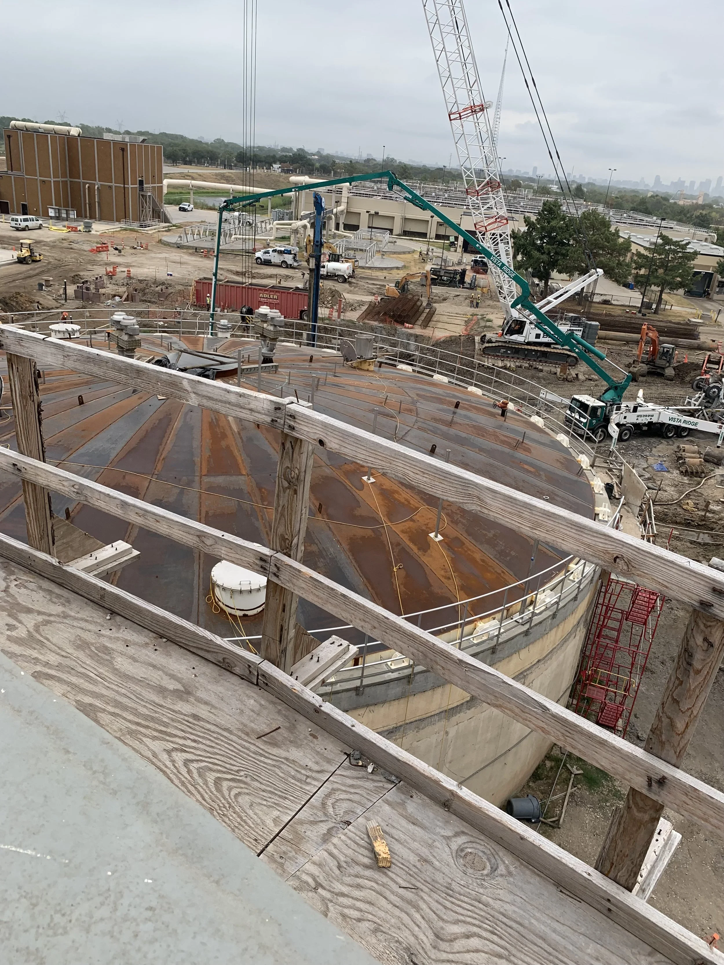 Construction site with a large rusty metal tank, cranes, trucks, and construction equipment.
