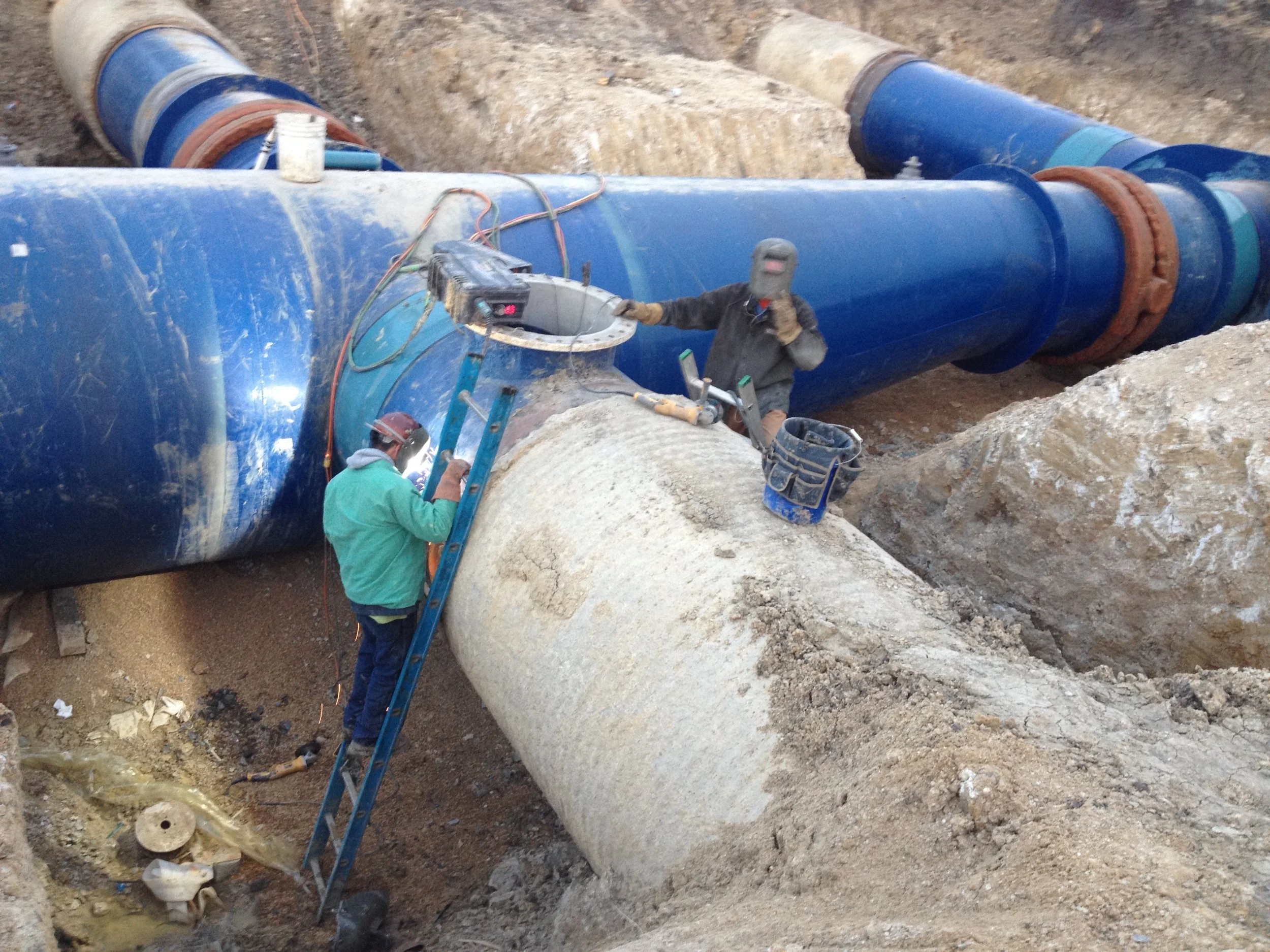 Two workers repairing large blue pipeline underground, with one on a ladder inspecting the pipe and another standing while talking on a phone.