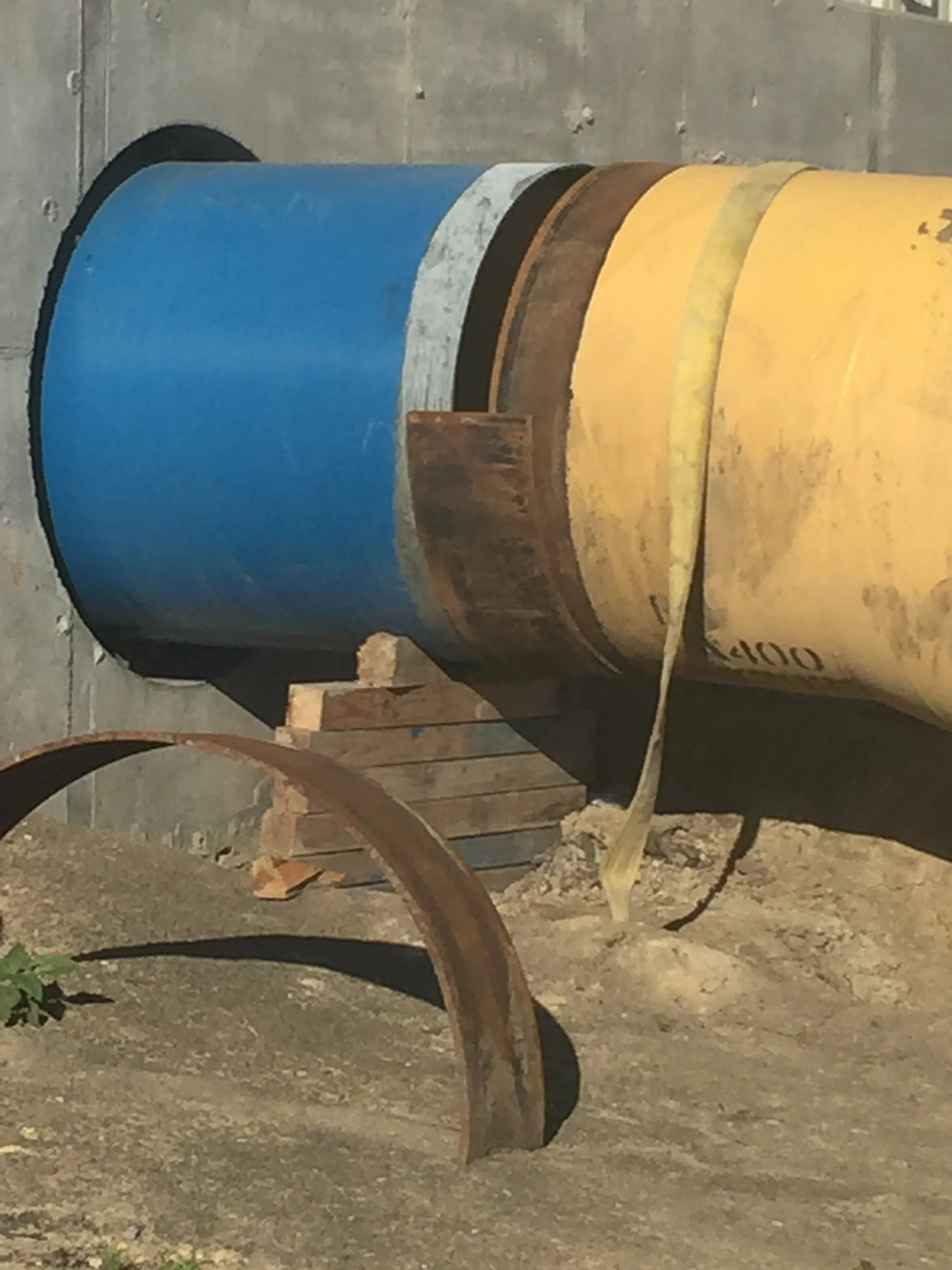 Two large rusty oil drums, one painted blue and the other yellow, are placed on the ground beside a wooden pallet and rusted metal pieces in an outdoor setting with a concrete wall in the background.