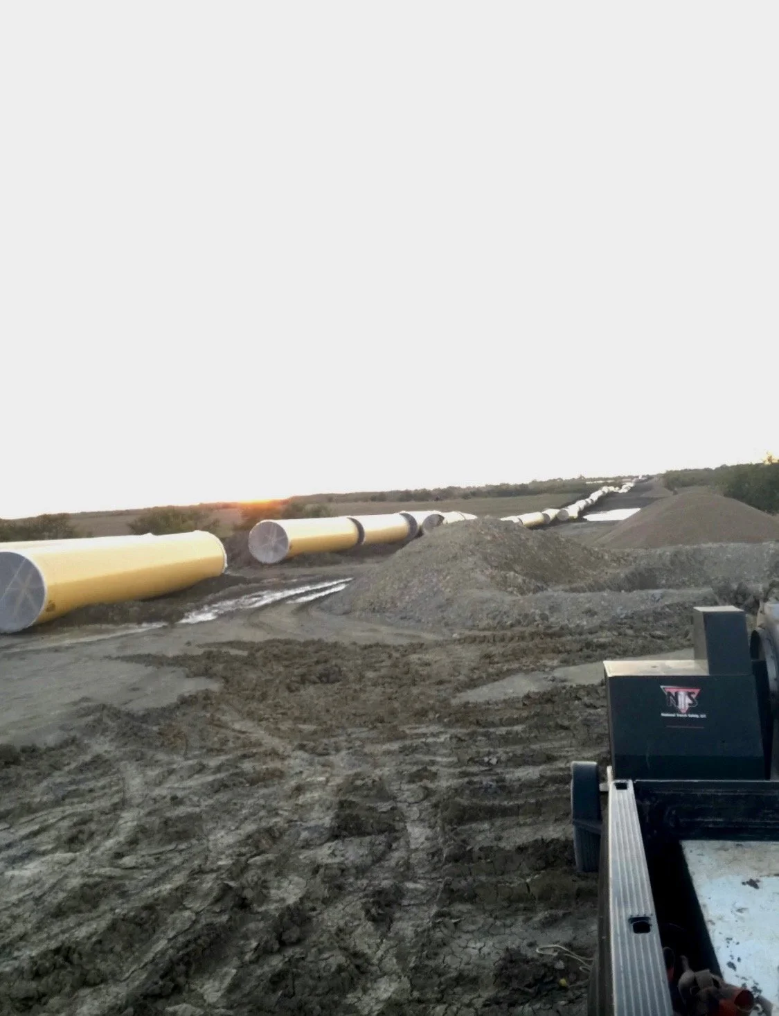 A construction site with yellow pipes laid out in a line on the ground, piles of dirt and gravel, and a piece of construction equipment on the right. The sunset is visible on the horizon.