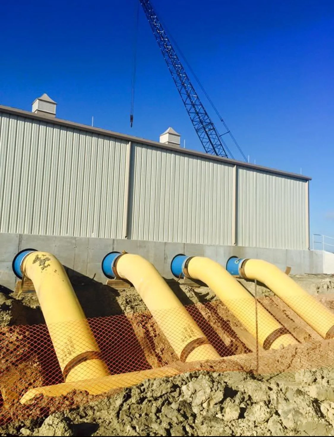 Construction site with large yellow pipes installed in dug earth, a beige metal building, and a blue crane against a clear blue sky.