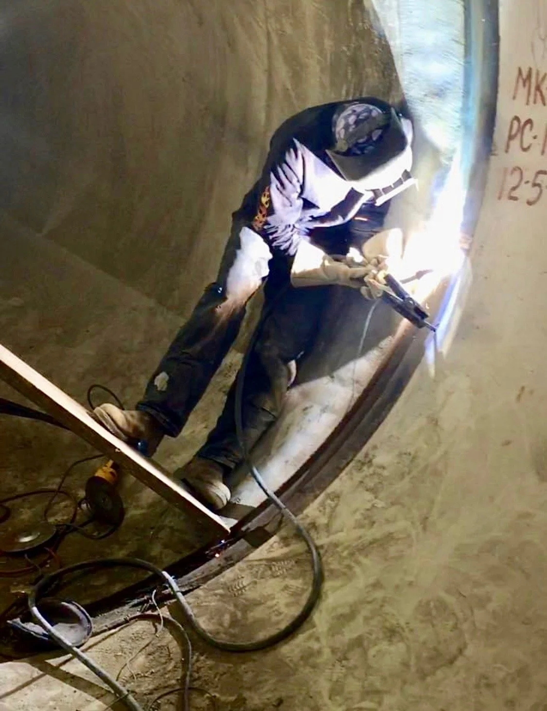 A person welding on a curved metal structure inside a construction site or workshop, with sparks and bright light from the welding process.