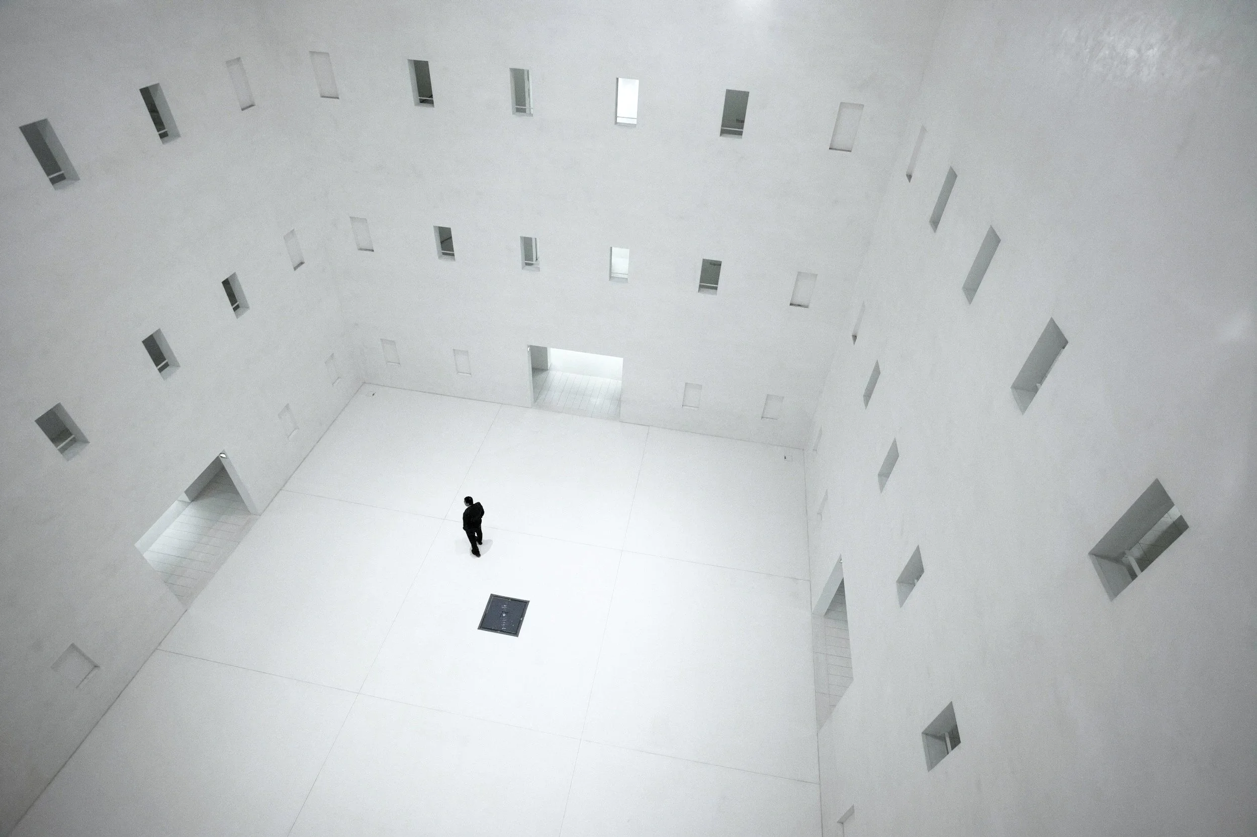 A person standing in a large, white, minimalist indoor space with multiple small rectangular windows.