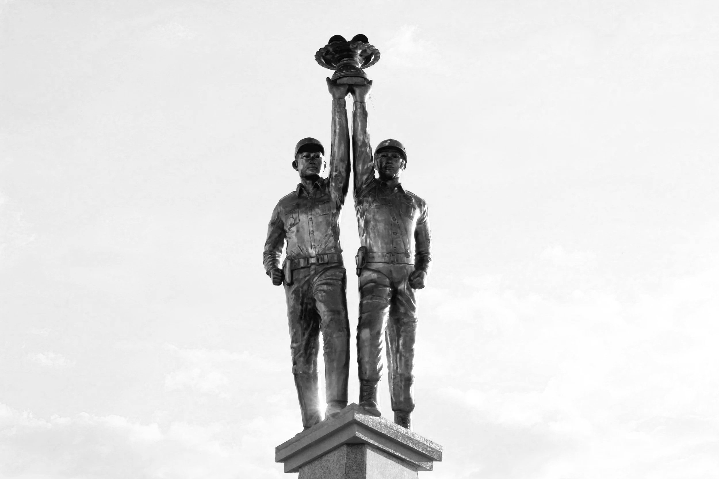 Black and white photo of two soldiers in uniform holding a wreath together above their heads, standing on a pedestal.