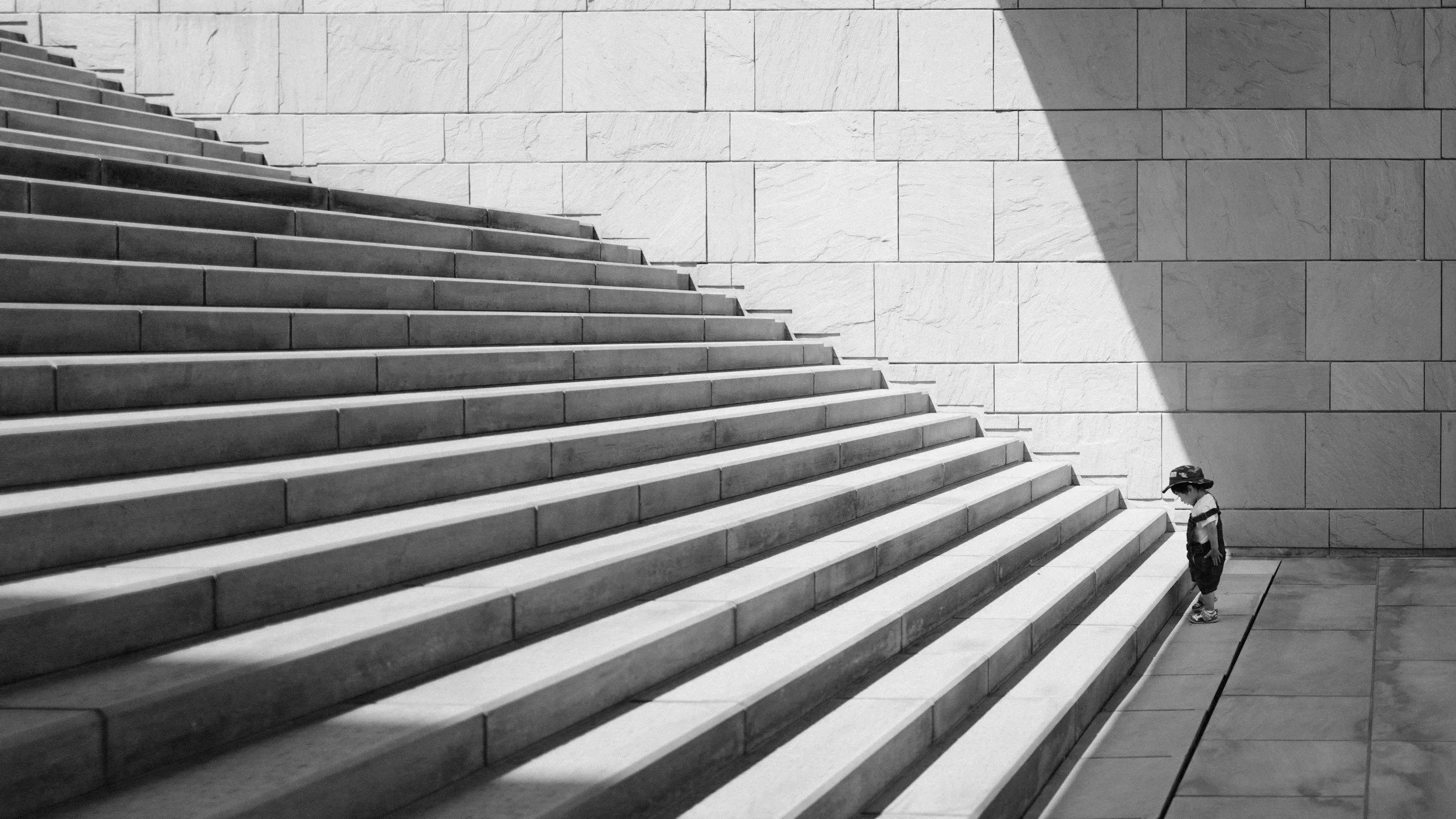 A young child with a hat standing at the bottom of a large, wide staircase made of stone, casting a shadow on the wall beside the stairs.