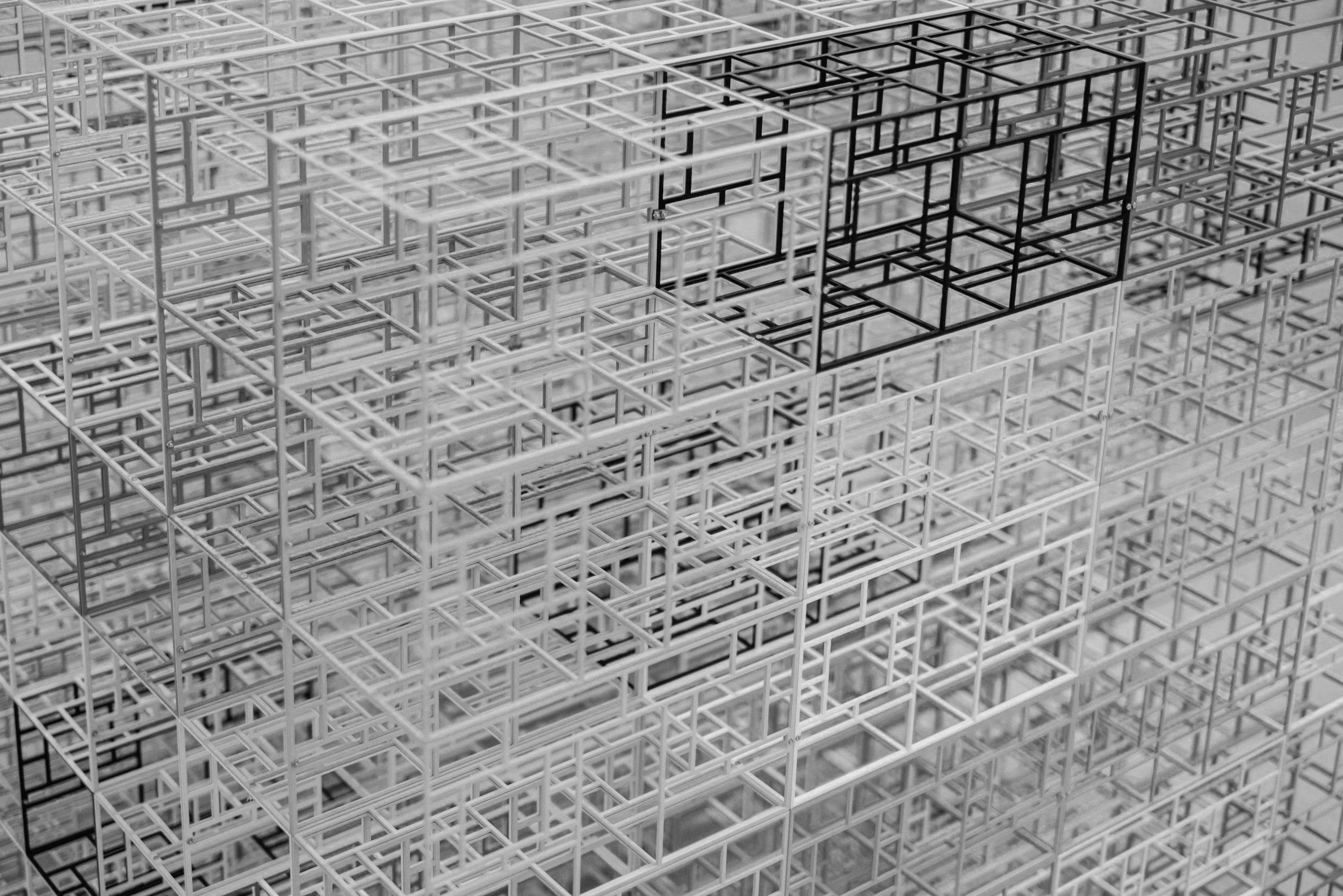 A close-up view of a 3D geometric structure made of interconnected metal or plastic frames in a grid pattern, with some frames colored black