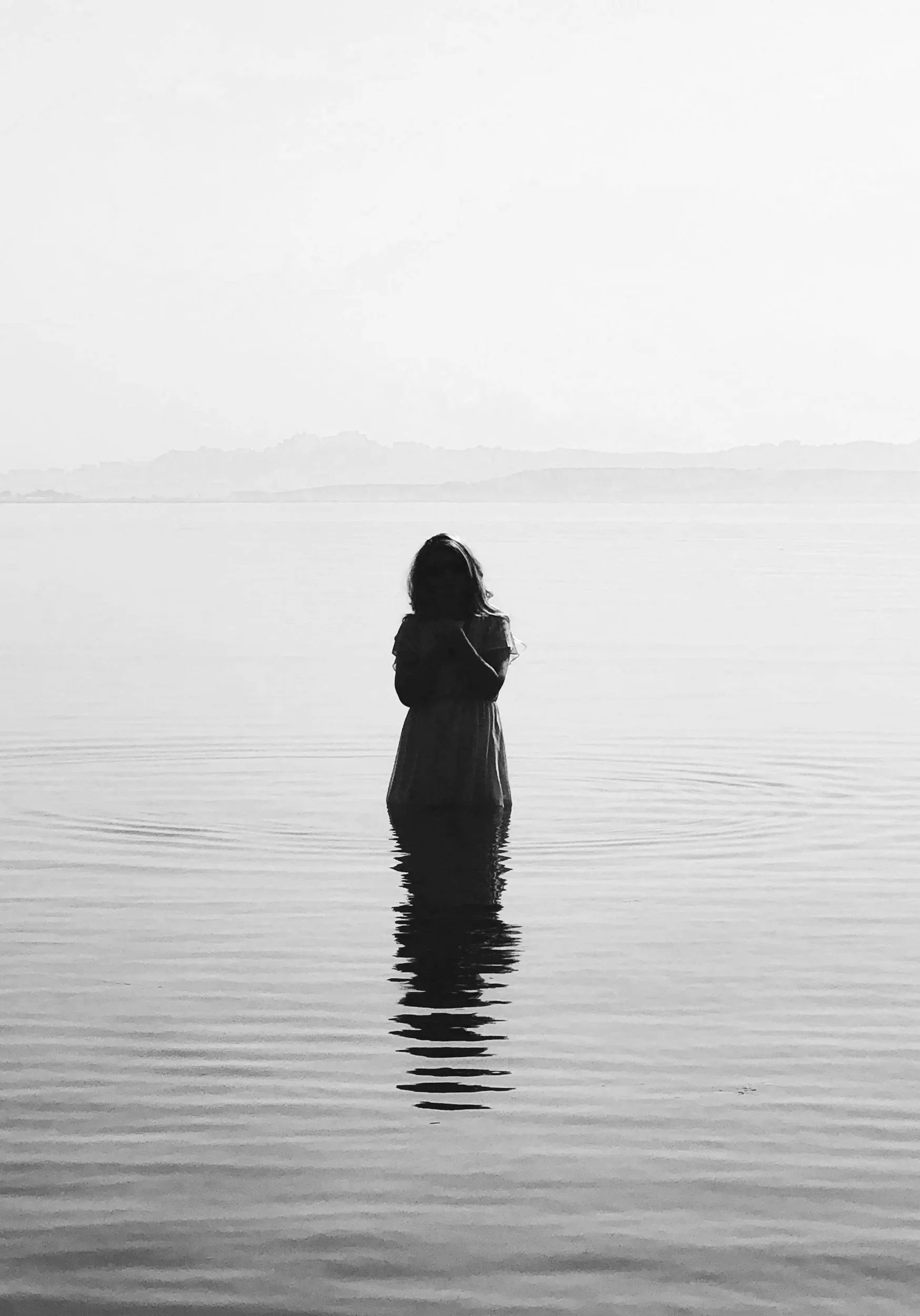 A person standing in water up to their waist, with long hair, facing away, against a calm body of water with distant land or mountains in the background, in black and white.