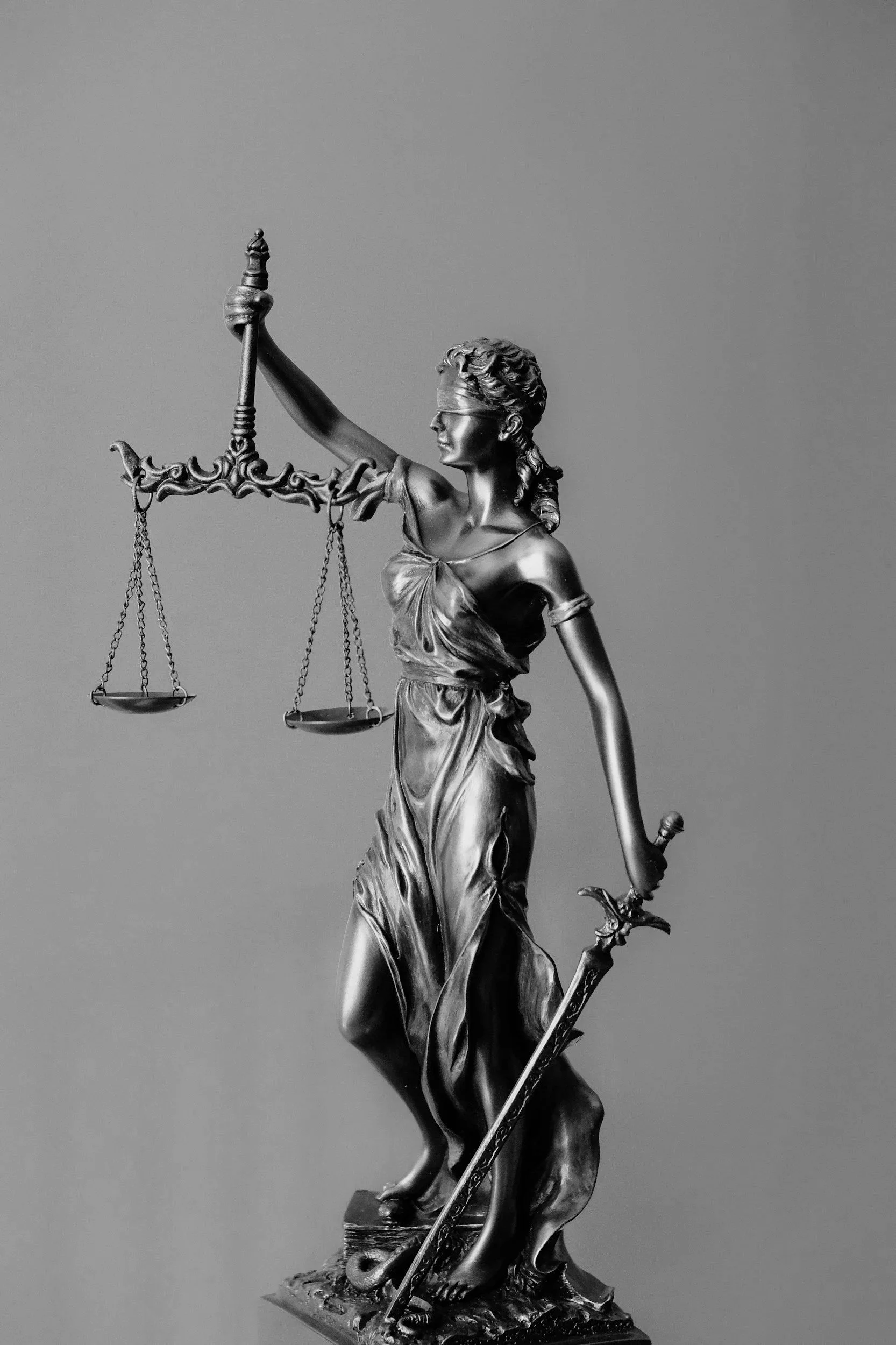 A black and white statue of Lady Justice holding a set of scales in her left hand and a sword in her right hand. She is blindfolded and dressed in flowing robes.