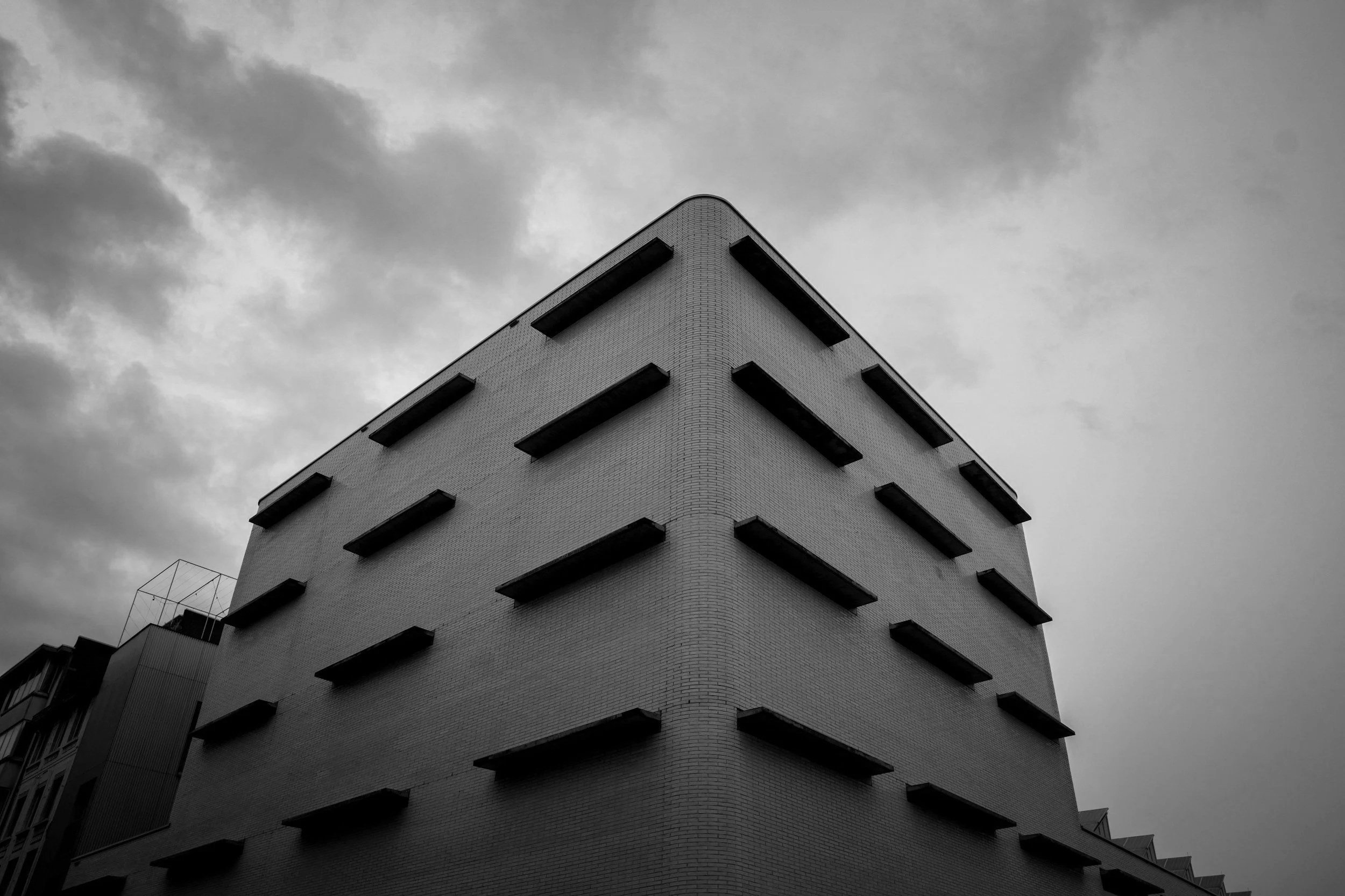 Black and white photo of a modern building with a pointed corner, multiple small rectangular windows, and cloudy sky in the background.