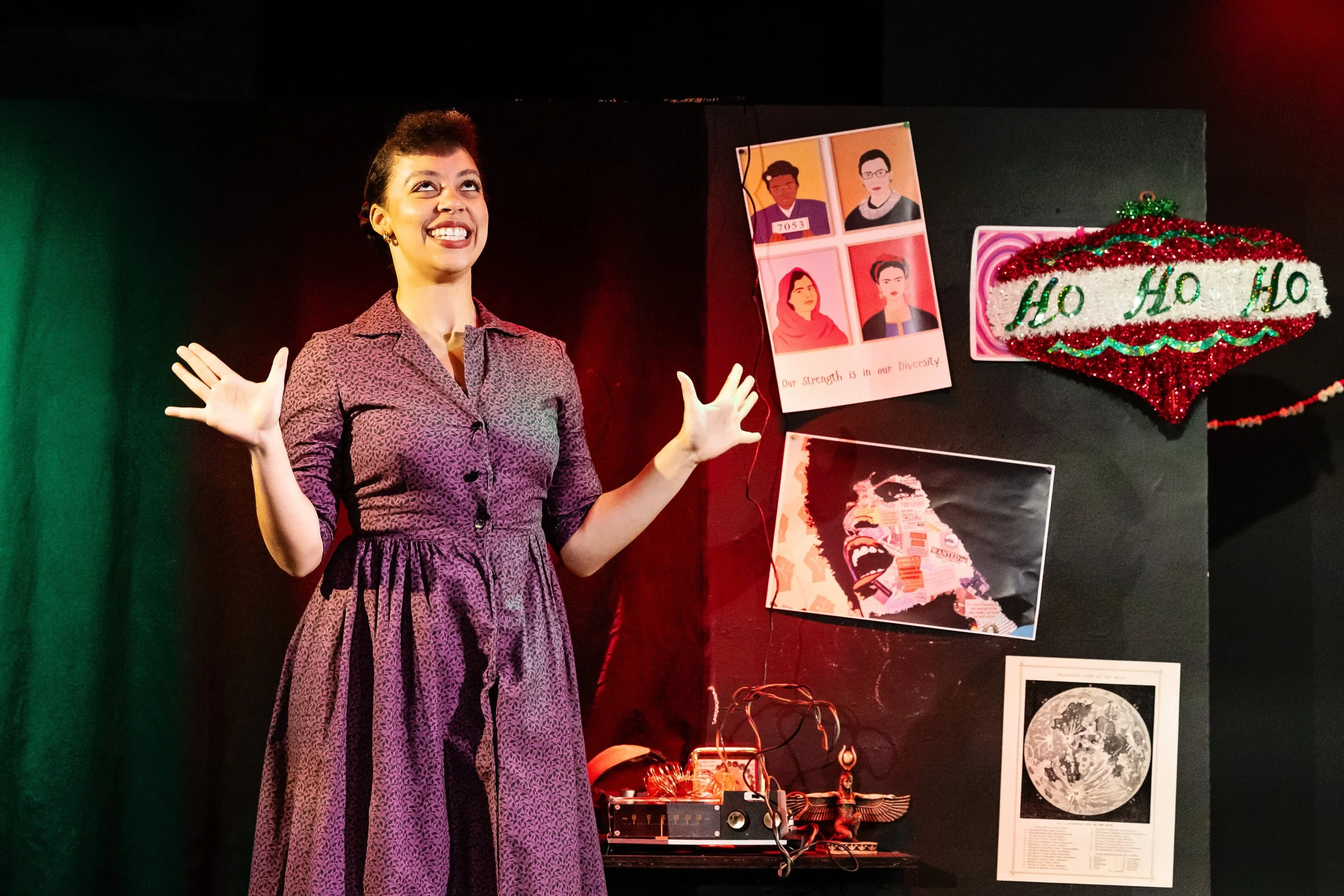 A woman standing on stage with a black background, smiling and gesturing with her hands. Behind her are posters and decorations, including a sign that says 'Ho Ho Ho' with red and green tinsel, and pictures of diverse people and artwork.