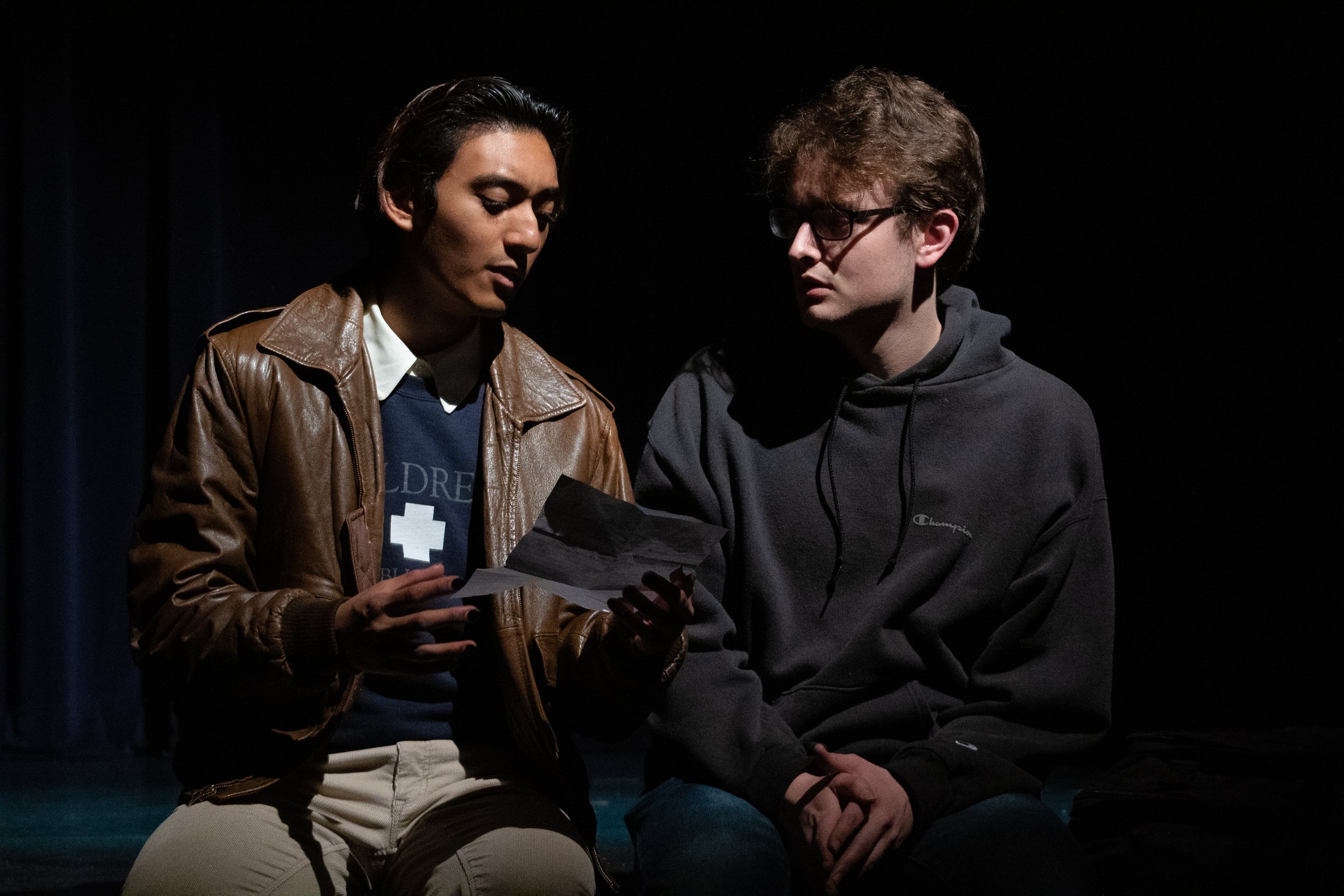 Two young men sitting and reading a piece of paper together in a dark setting.