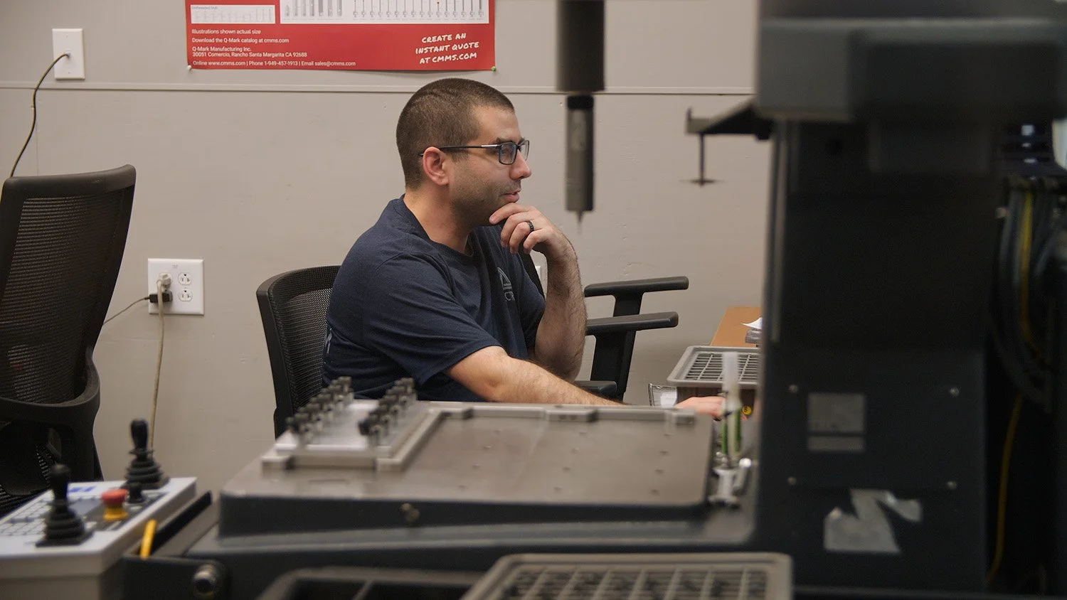 A man with short hair, glasses, and a beard, sitting at a desk in an office, working on a computer. He is wearing a dark t-shirt and looking at the monitor with his hand on his chin. The workspace has equipment and a poster on the wall behind him.