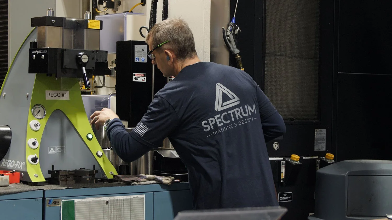 aerospace precision machining A man working in a machine shop setting, wearing safety glasses, a headset, and a lab coat with the logo 'Spectrum Machine & Design.' He is operating or inspecting a green and gray piece of industrial equipment