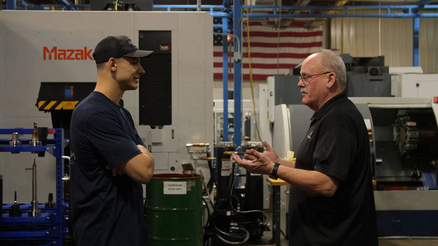 aerospace precision machining Two men are having a discussion in an industrial factory with machinery in the background. One is younger, wearing a black cap and navy shirt, and the other is older with glasses, wearing a black polo shirt.