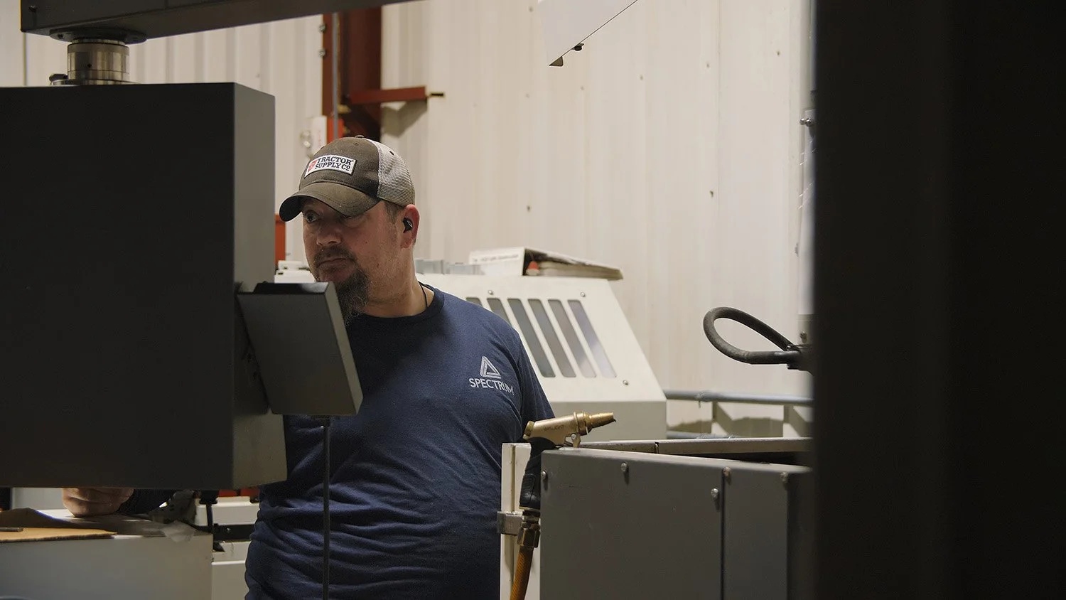 aerospace machinist jobs Ohio A man wearing a baseball cap and a blue T-shirt with 'SPECTRUM' on it, working with industrial machinery in a workshop or factory.