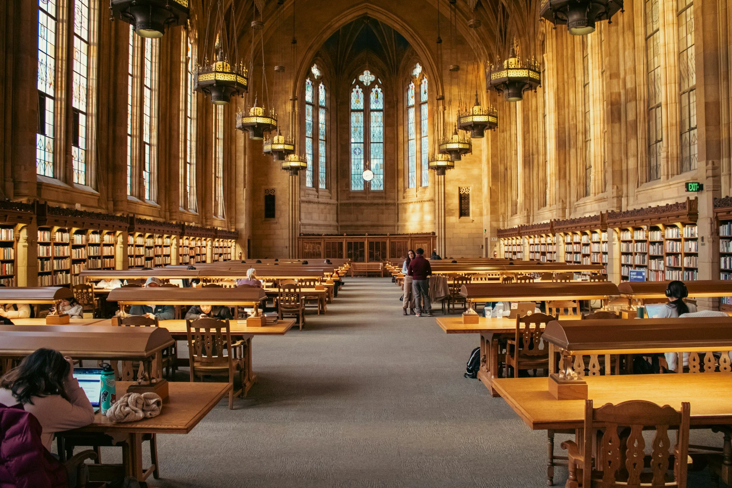 Interior of a historic library with tall stained glass windows, wooden bookshelves, and long study tables with people reading and working. Adjunct Faculty. Counselor Education.