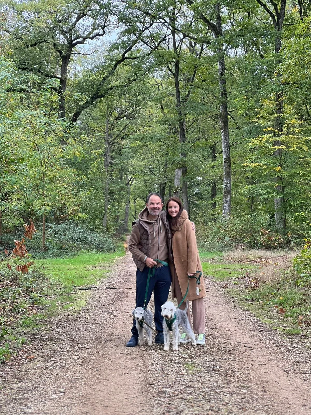 A smiling man and woman standing on a dirt trail in a wooded area, each holding a leash with a dog, surrounded by green trees and foliage.