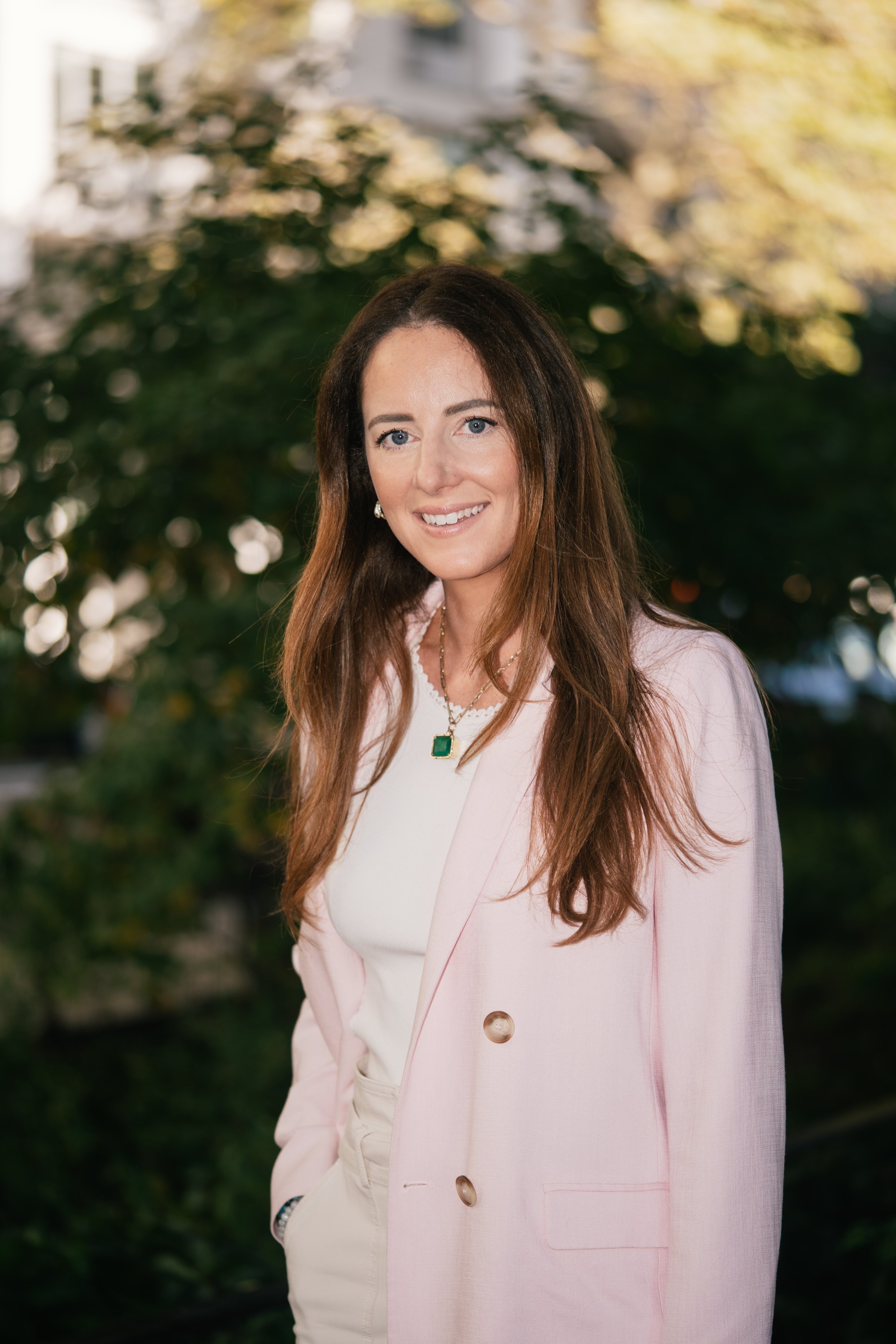 A woman with long brown hair smiling outdoors, wearing a light pink blazer over a white top and adorned with green and gold jewelry, with blurred greenery in the background.