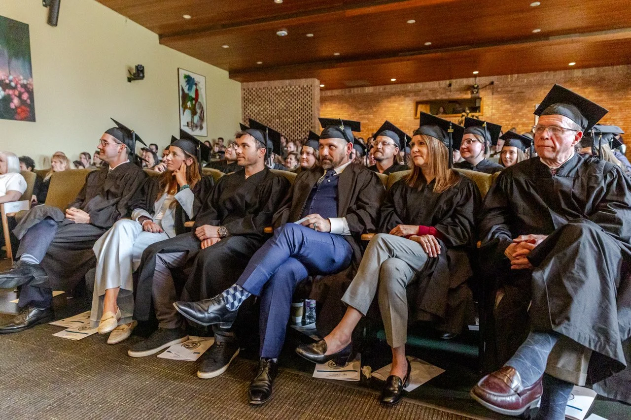 A group of graduates seated in an auditorium during their graduation ceremony, wearing black caps and gowns.