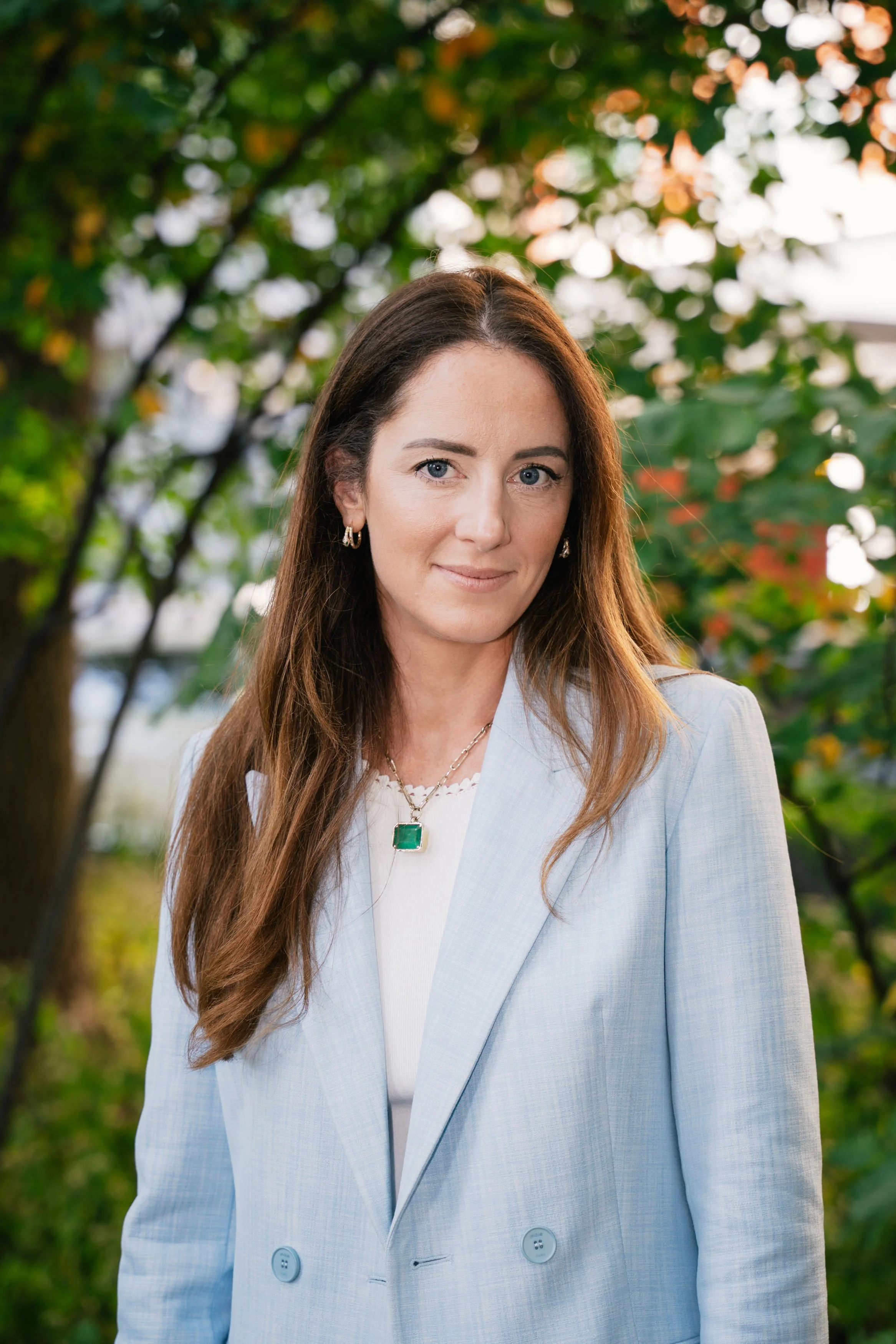 A woman with long brown hair, blue eyes, and light skin, wearing a light blue blazer and a necklace with a green pendant, standing outdoors with trees and foliage in the background.