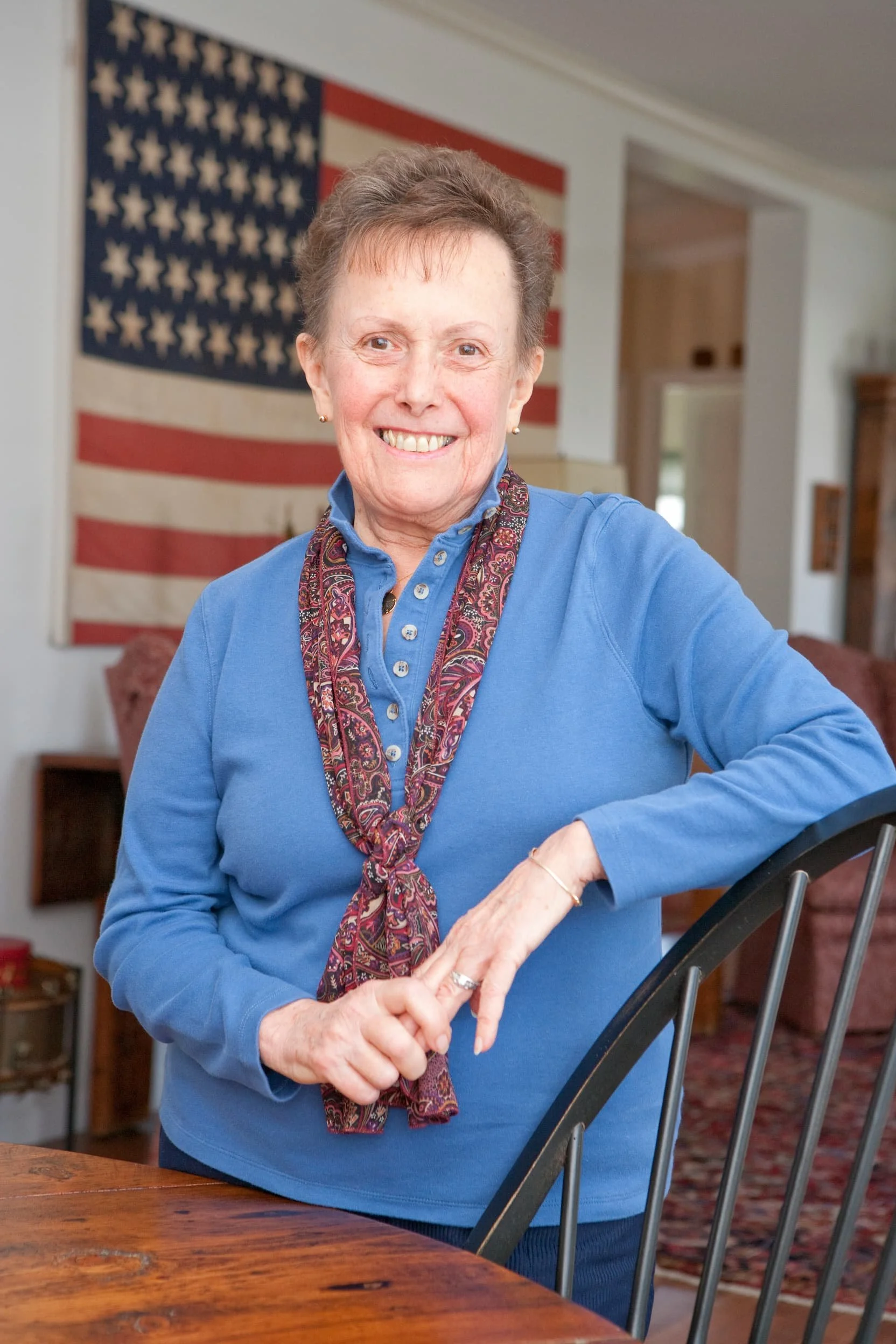 An elderly woman with short hair, smiling, wearing a blue sweater and a patterned scarf, standing indoors in front of an American flag. She is resting her left hand on a black metal chair and standing next to a wooden table.