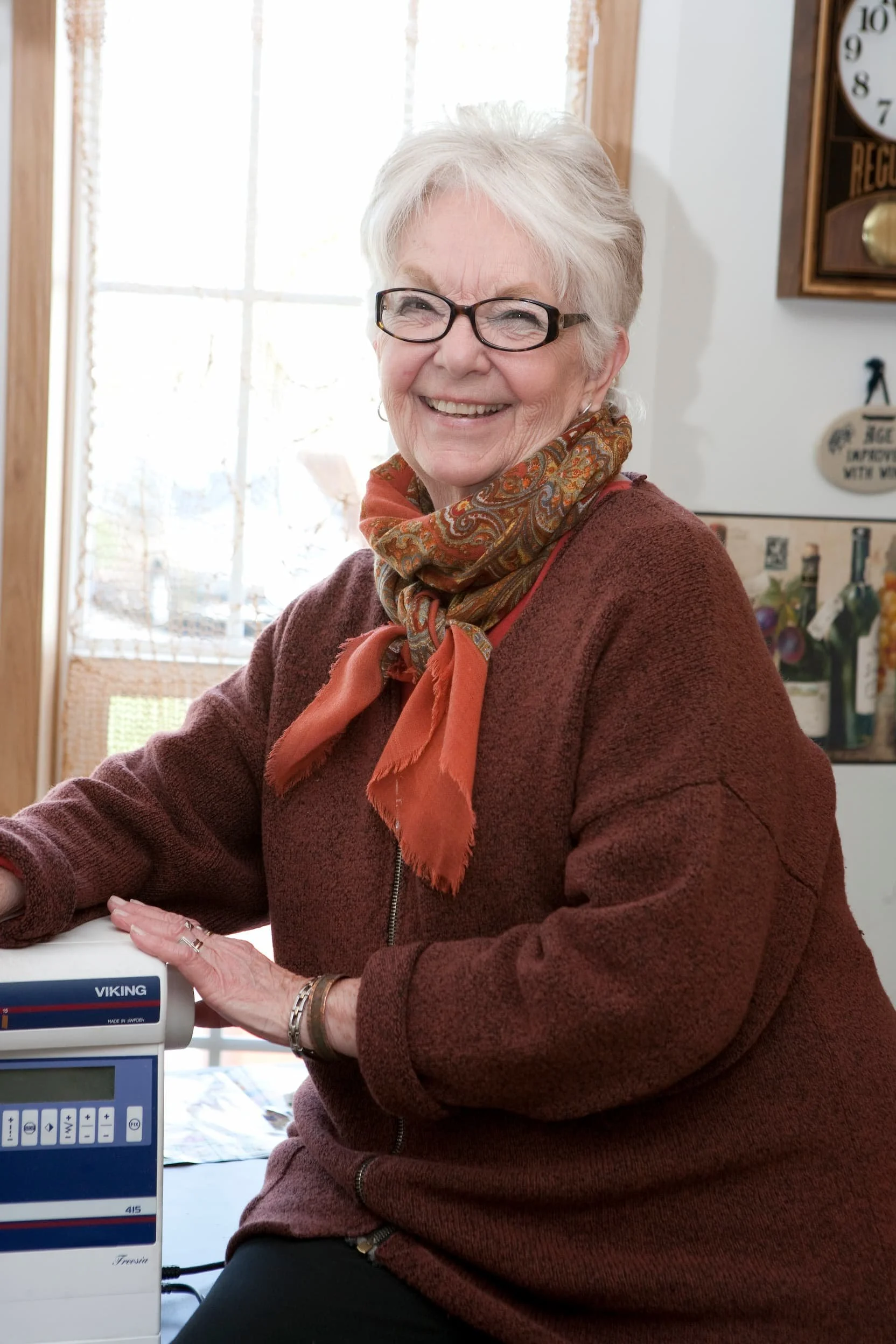 An elderly woman with gray hair, glasses, and a warm smile sitting indoors near a window, wearing a patterned scarf and a maroon sweater.