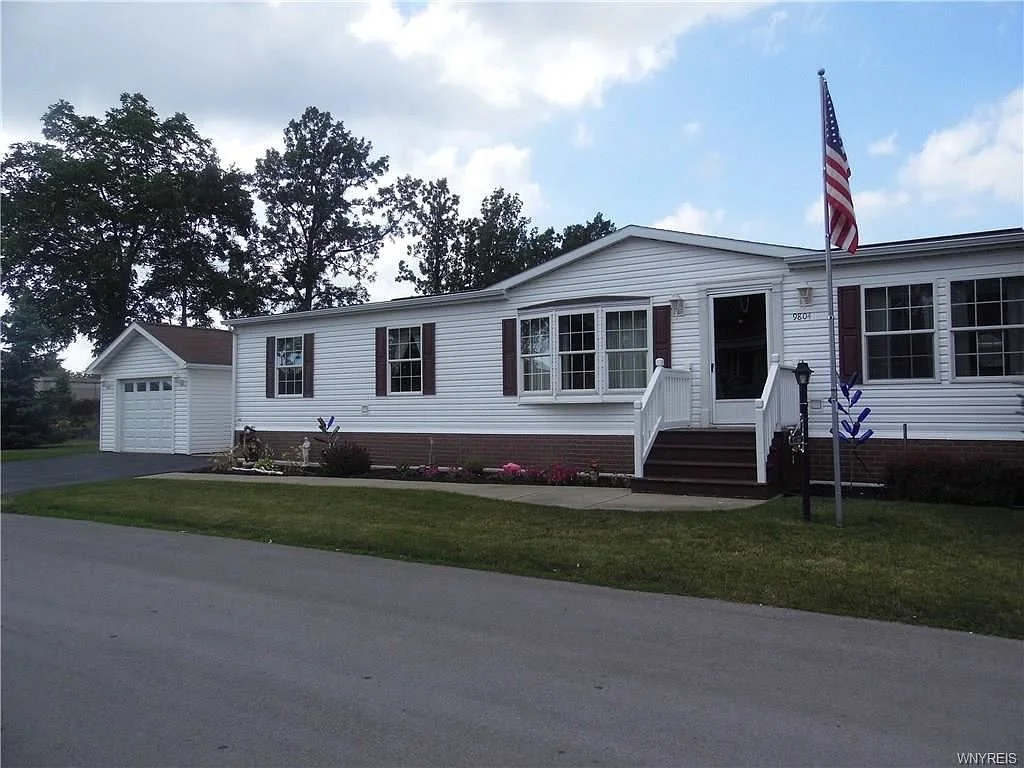 White manufactured home with brown shutters, front porch, path, American flag, greenery, and a small garage, under a partly cloudy sky.