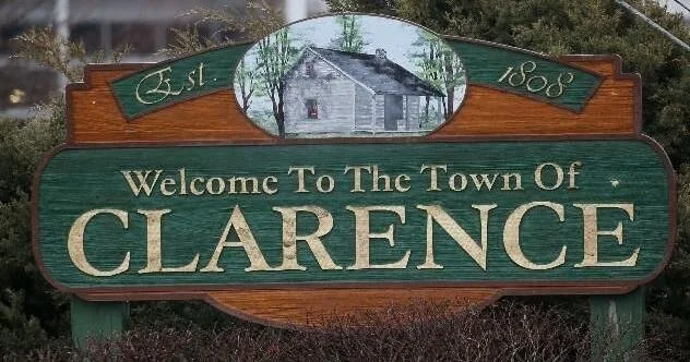 A wooden welcome sign for the town of Clarence, Vermont, established in 1800, featuring a painted image of a house and trees.