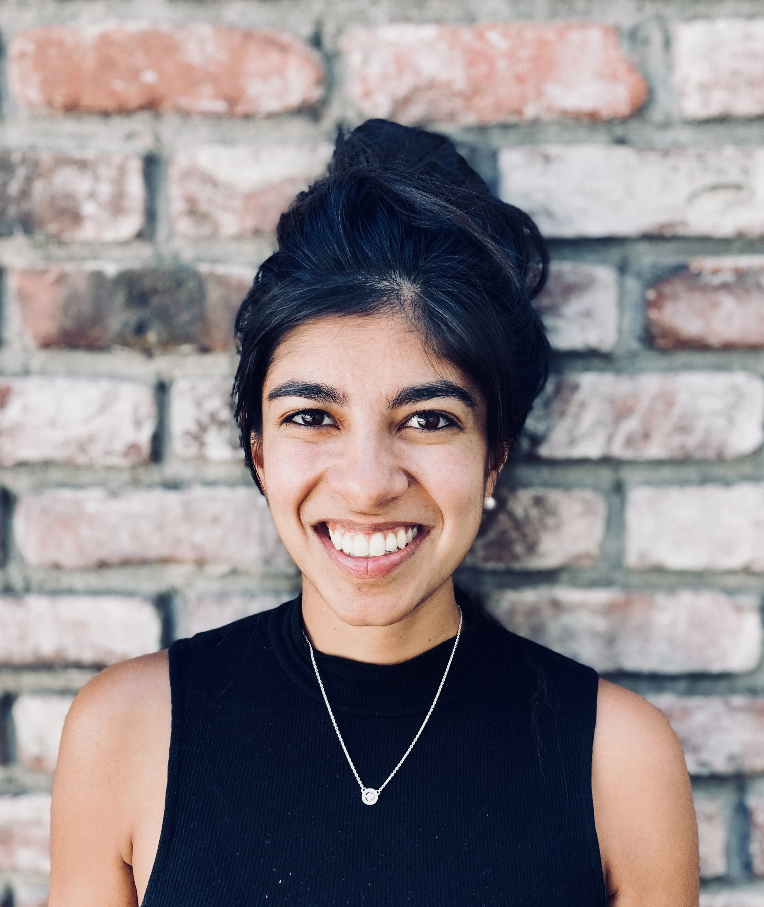 A young woman with short black hair and a big smile, wearing a black sleeveless top and a necklace, standing against a brick wall.