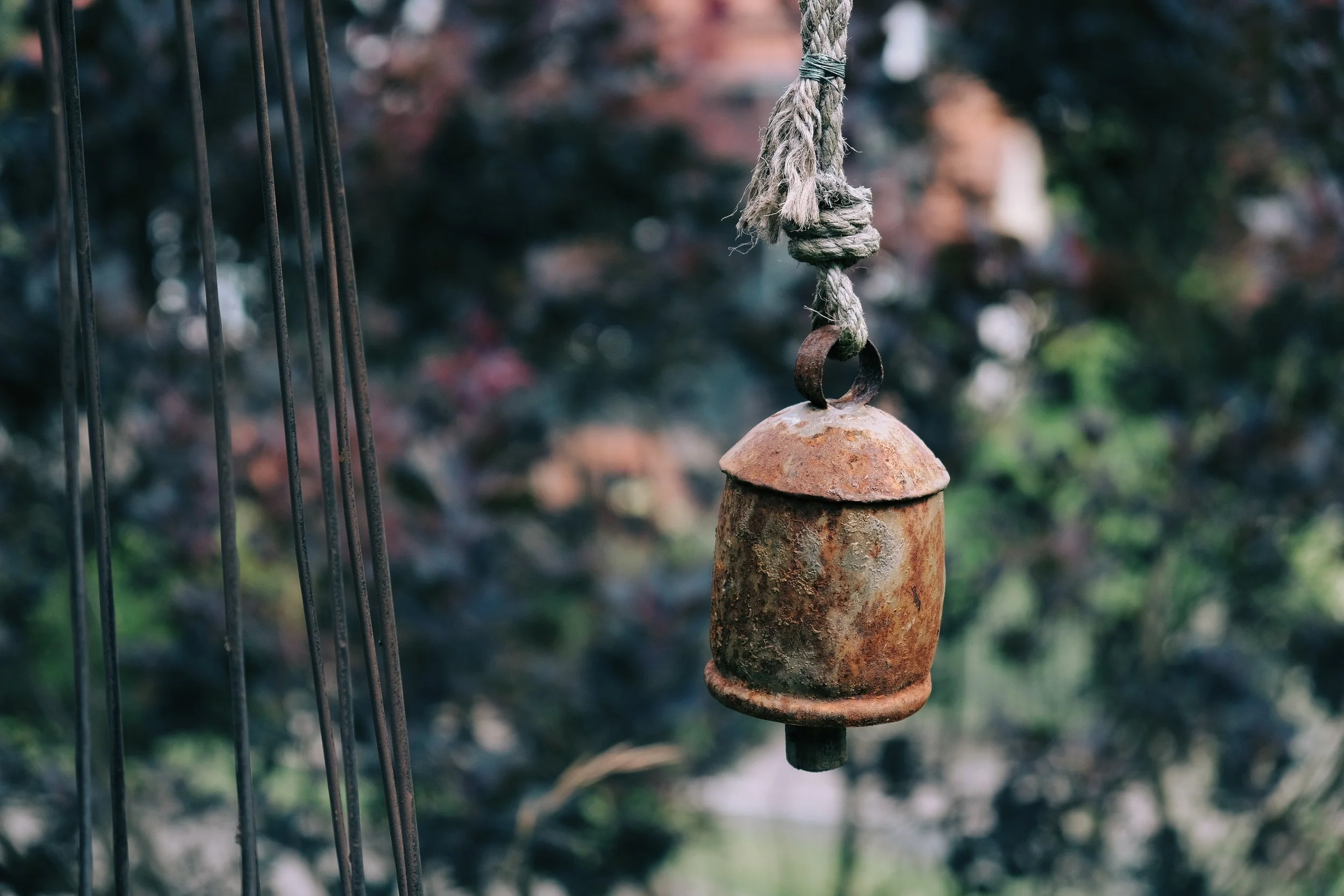 An old, rusted hanging bell made of metal, suspended by a frayed rope, outdoors with a blurred background of trees and foliage.