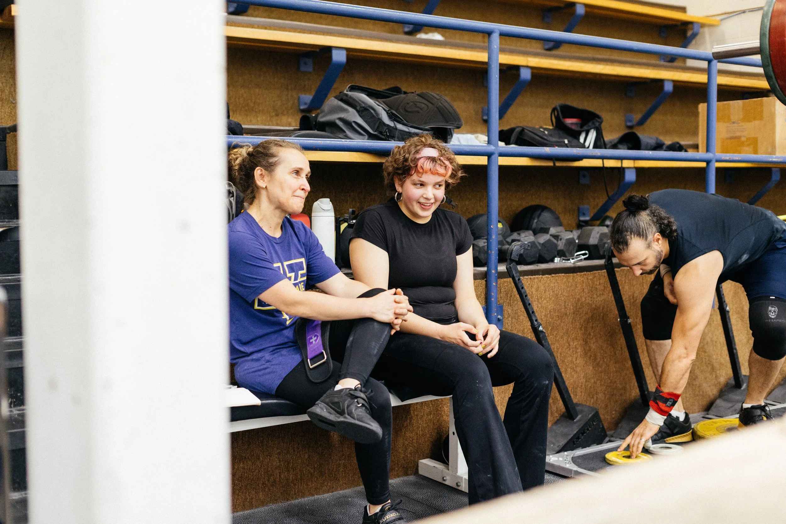 Groupe de trois personnes dans une salle de sport ou salle d'entraînement, avec des haltères et des sacs de sport en arrière-plan. Deux femmes assises discutent, une homme enfilant des chaussures de sport.