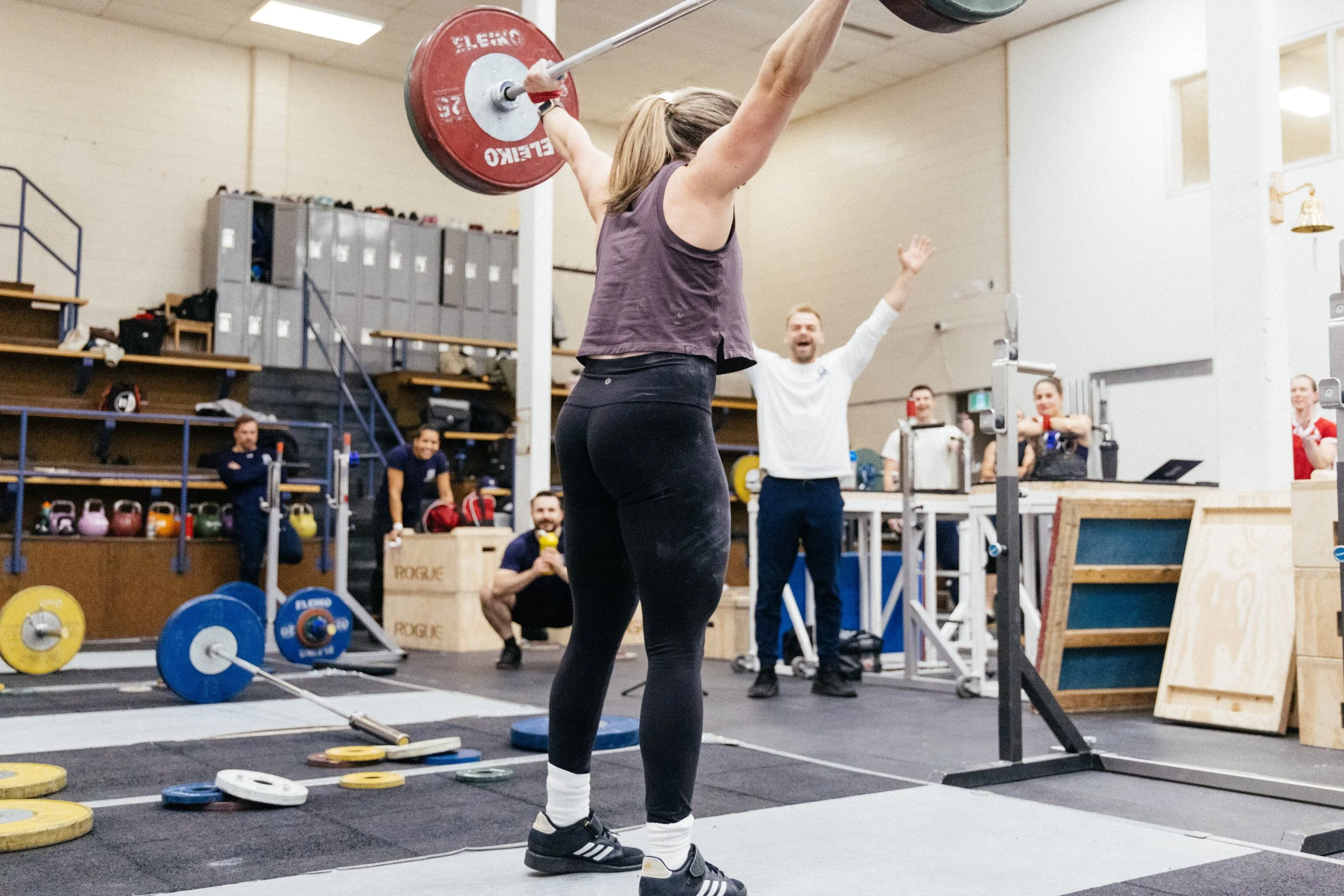 Une femme en tenue sportive soulève une barre avec des poids en salle de musculation, entourée de personnes qui regardent et encouragent, dans un espace équipé pour l'entraînement.