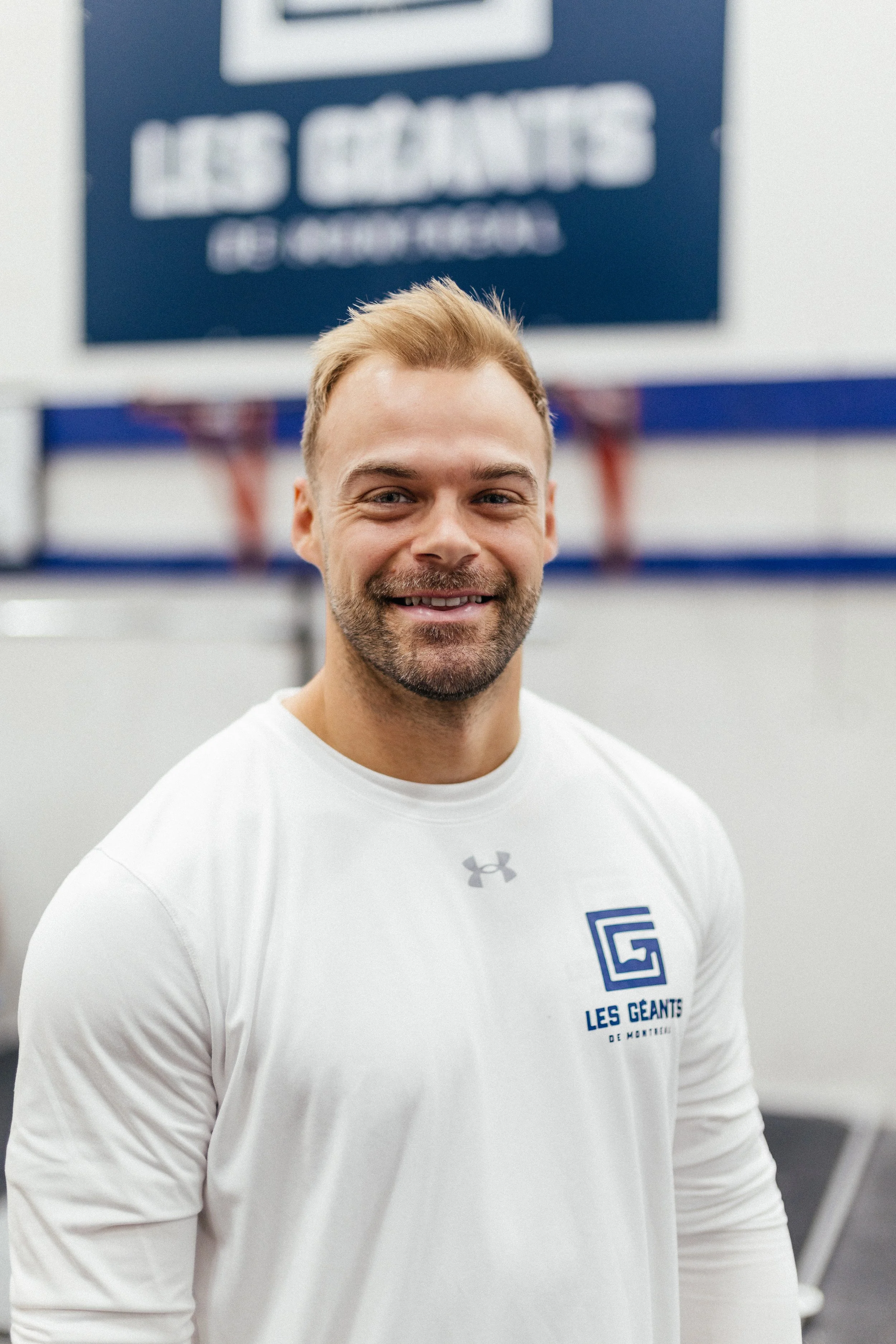 Un homme souriant portant un t-shirt blanc avec le logo et le texte 'Les Geants de Montaigu', dans un environnement intérieur
