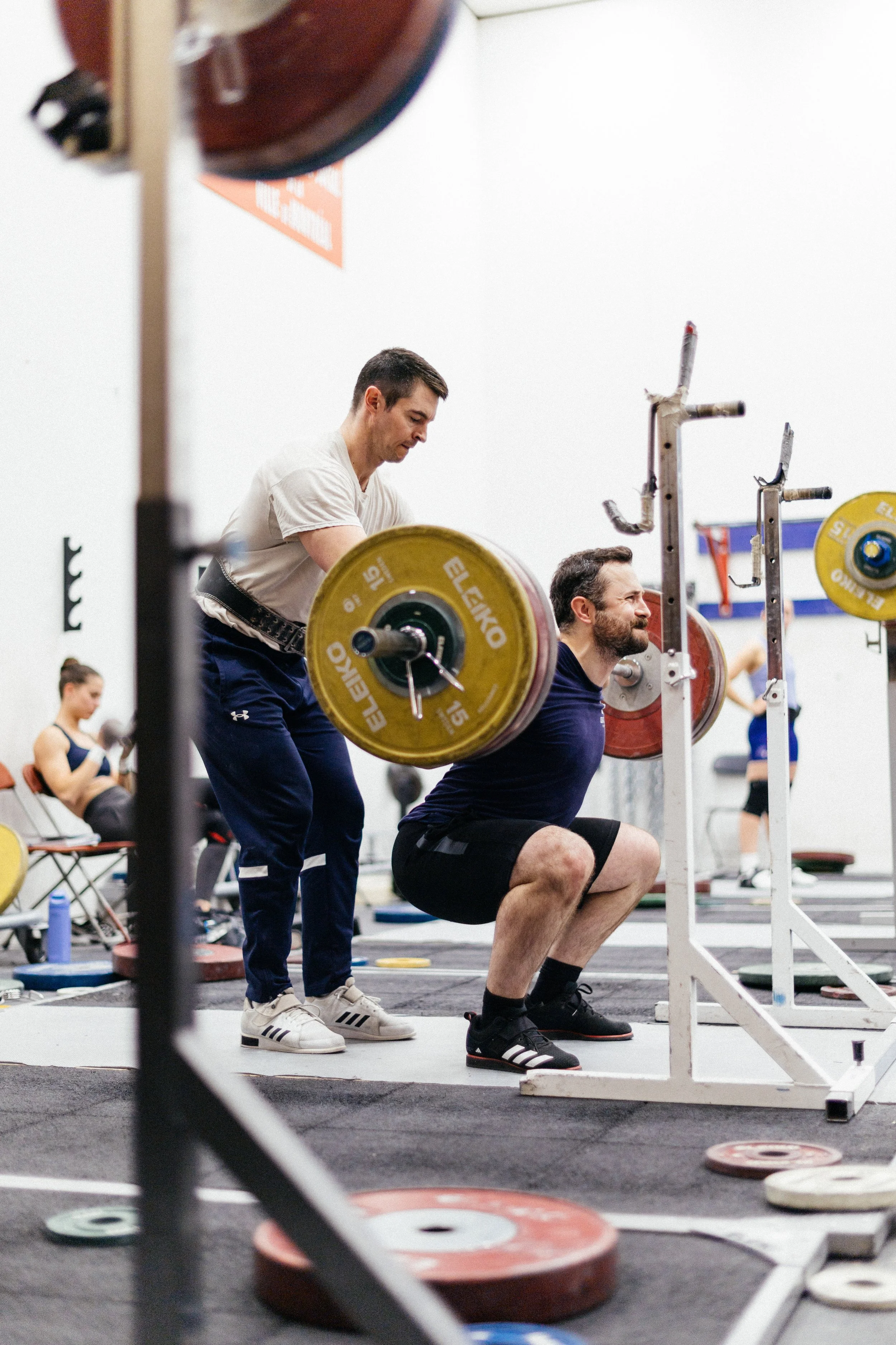Homme en train de faire un squat avec une barre chargée de poids dans une salle de sport, superviseur à ses côtés, autres personnes en arrière-plan.