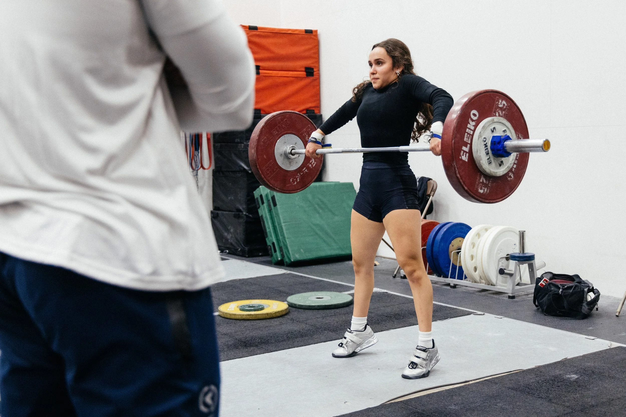 Une femme en train de faire de l'haltérophilie dans une salle de sport, ne portant qu'un t-shirt noir, un short noir, et des chaussures de sport, entourée de plaques de poids colorées.