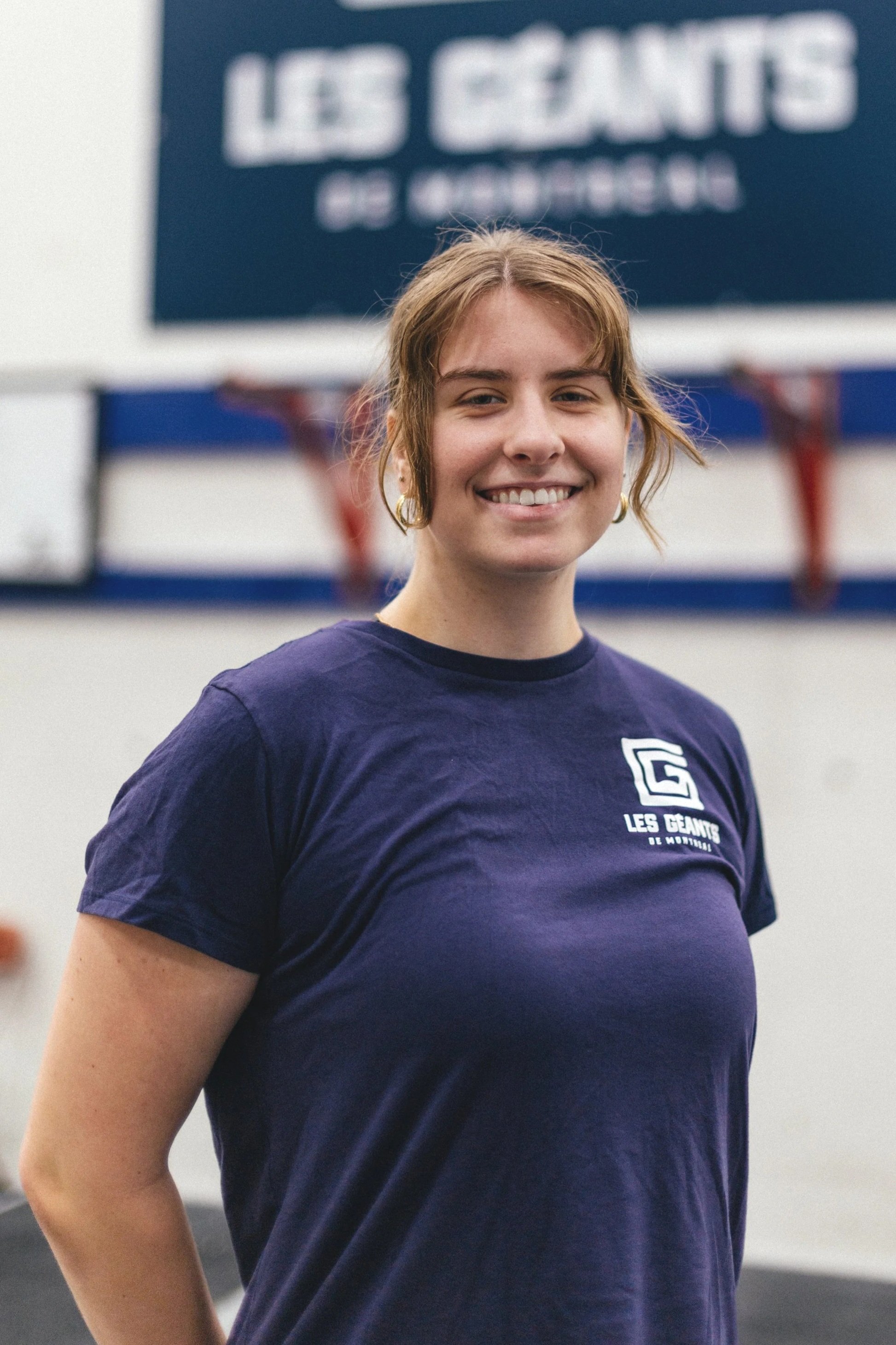 Jeune femme souriante portant un T-shirt bleu avec un logo et le texte "Les Géants de Montréal", dans un environnement intérieur.