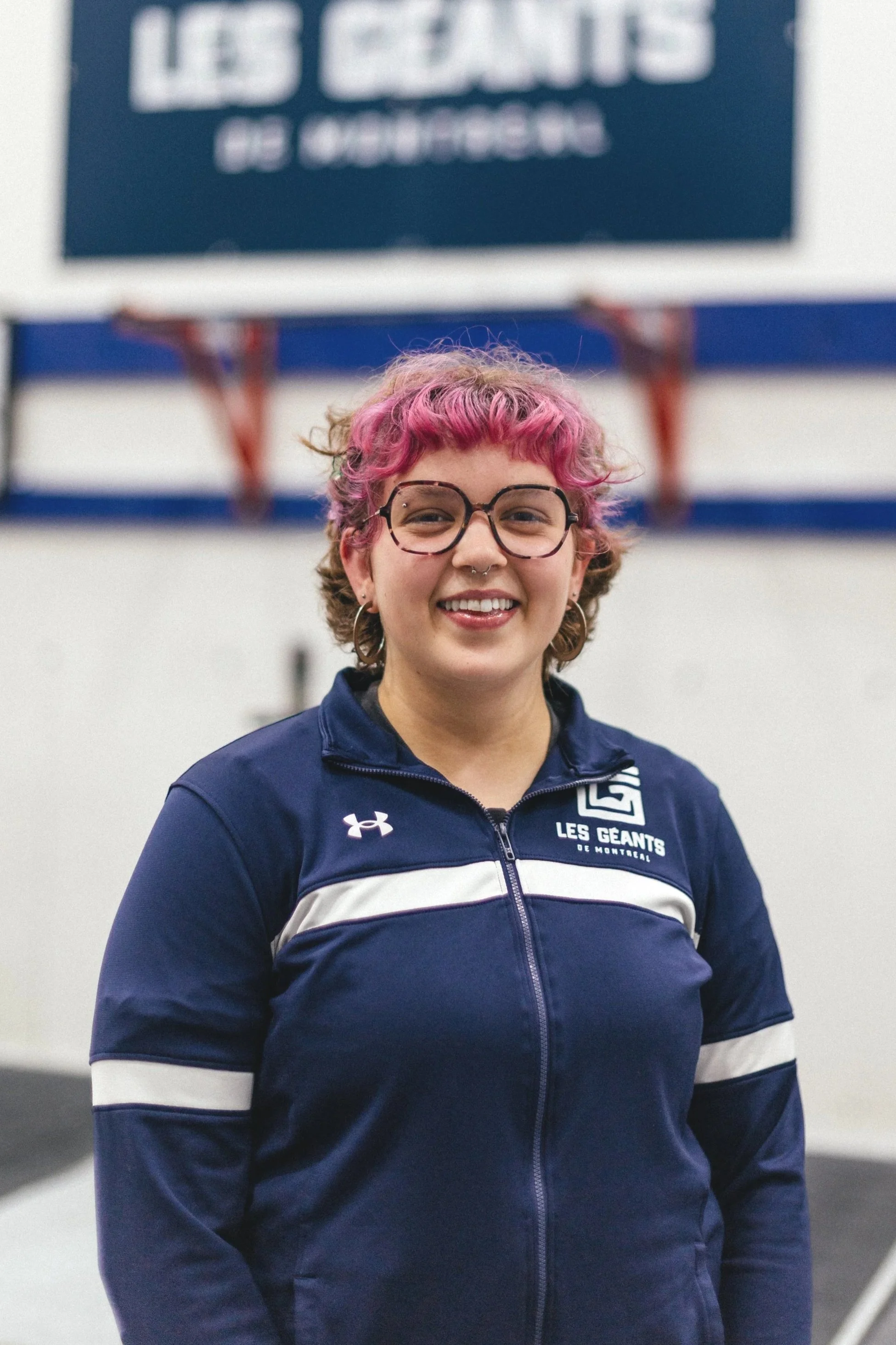 Une jeune femme avec des cheveux bouclés et teints en rose, portant des lunettes rondes, un sweat à capuche de l'équipe Les Géants de Montréal, souriante en posant à l'intérieur d'une salle.